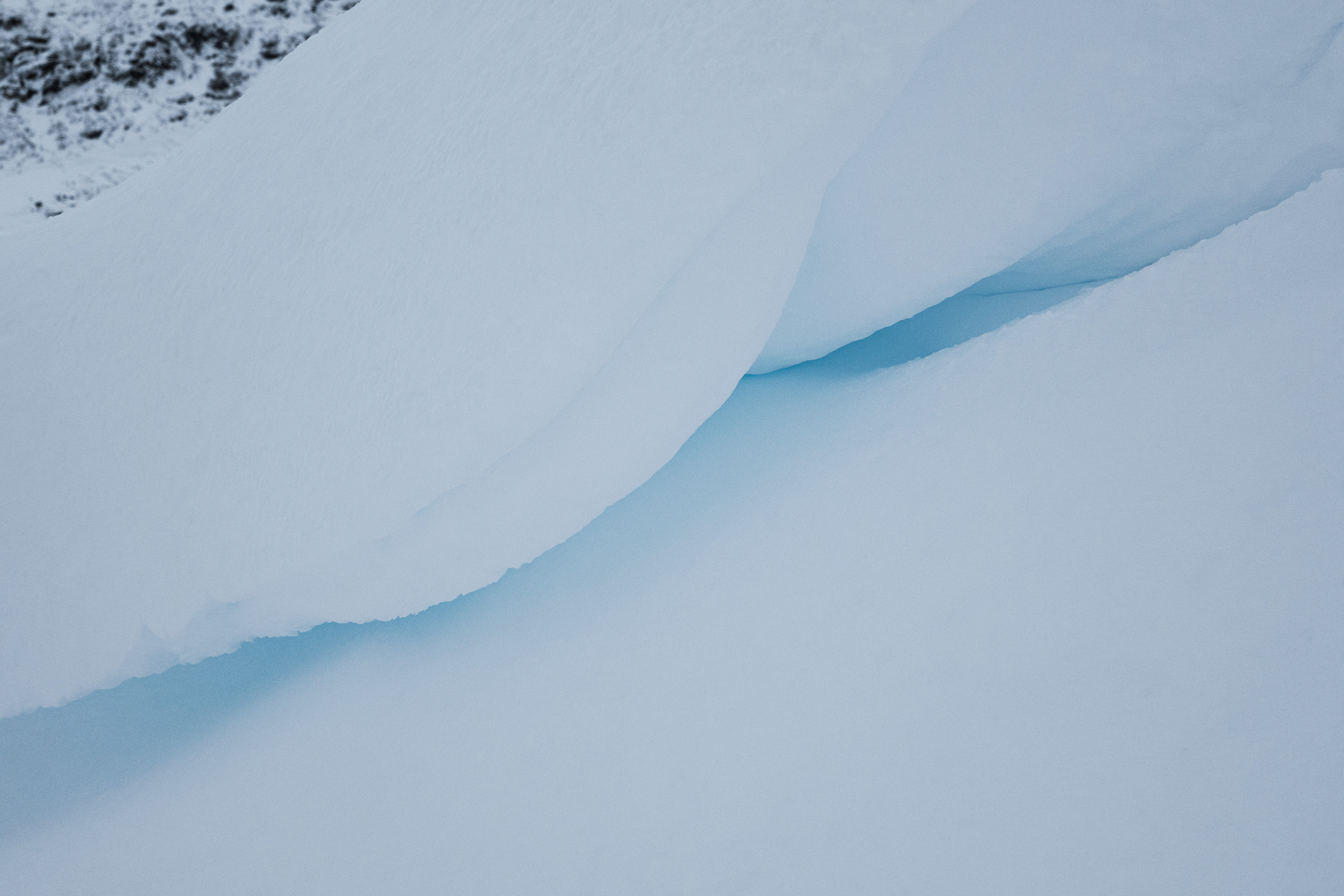 Close-up image of overlapping snow and ice layers forming a naturally sculpted, wave-like ridge with a soft blue gradient in the center, suggesting the presence of compacted glacial ice. The upper left corner reveals a hint of a dark, rocky terrain, providing contrast to the smooth, cold surfaces.