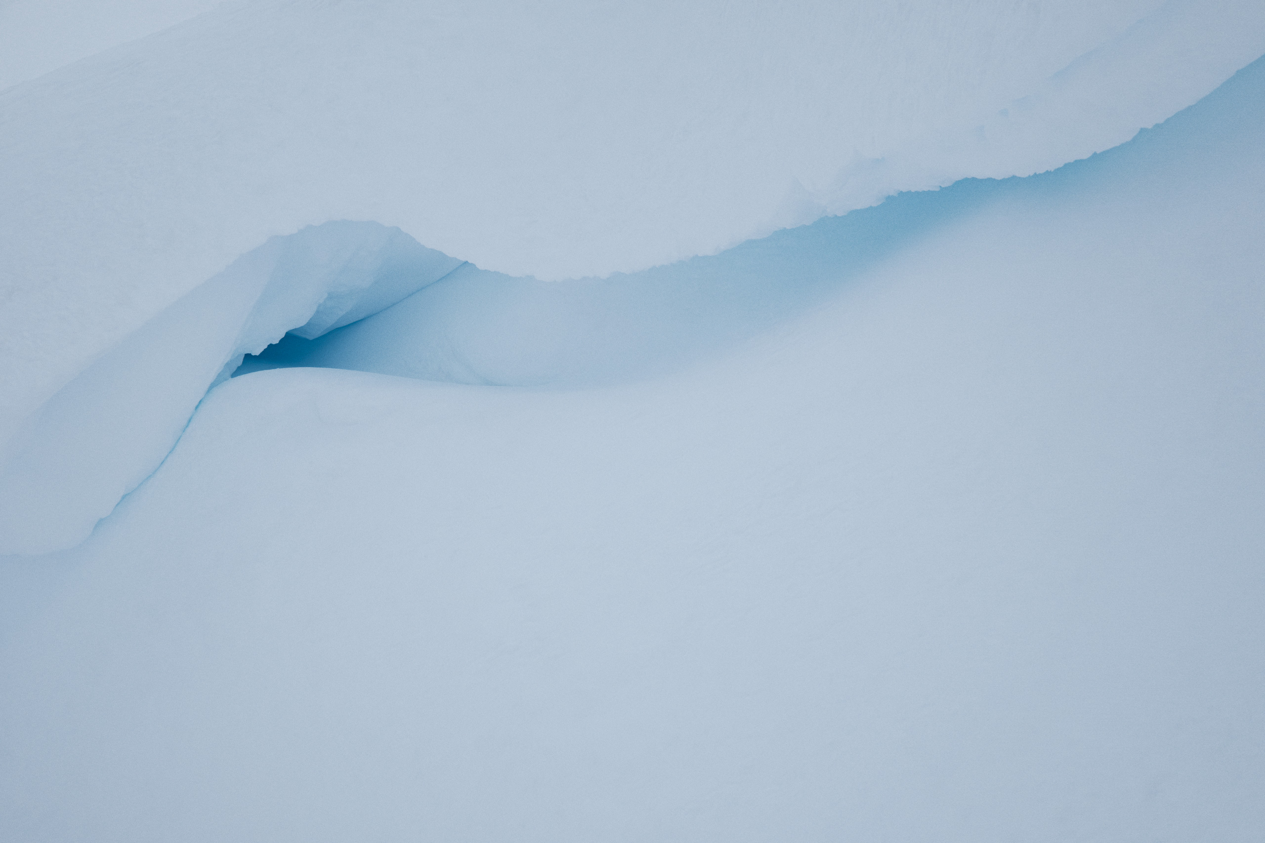 Minimalist close-up image of a smooth snowbank with a natural, wave-like indentation revealing a layer of dense, bluish glacial ice beneath the surface. The gentle curvature and soft gradient from white to icy blue emphasize the organic form and texture of the frozen landscape.