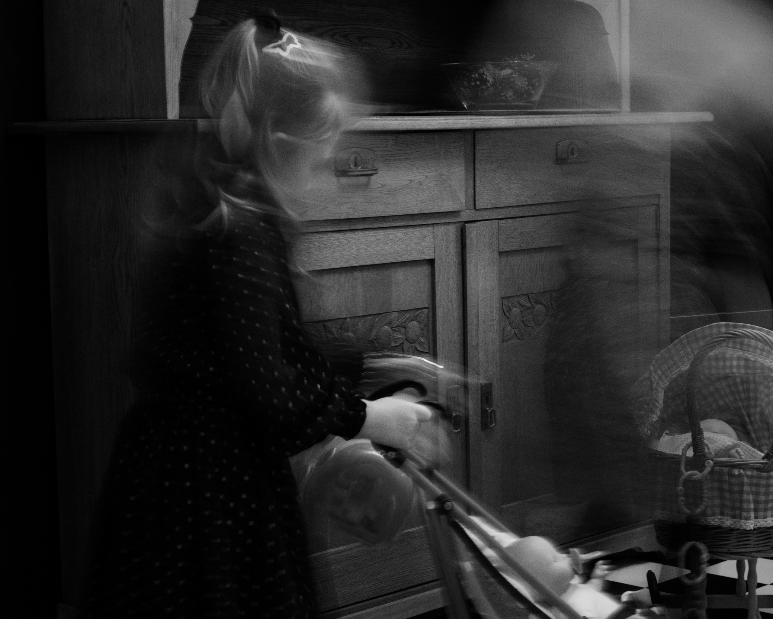 Black and white photo of a young girl with long blonde hair in a dark polka-dot dress pushing a vintage-style baby stroller in front of a carved wooden cabinet. The image has a ghostly, double exposure or long-exposure blur effect, creating a dreamlike sense of movement. A doll’s head is visible in the stroller, and a checkered blanket or cloth partially covers another doll or toy. The cabinet behind features detailed floral wood carvings and a bowl with decorative items on top.