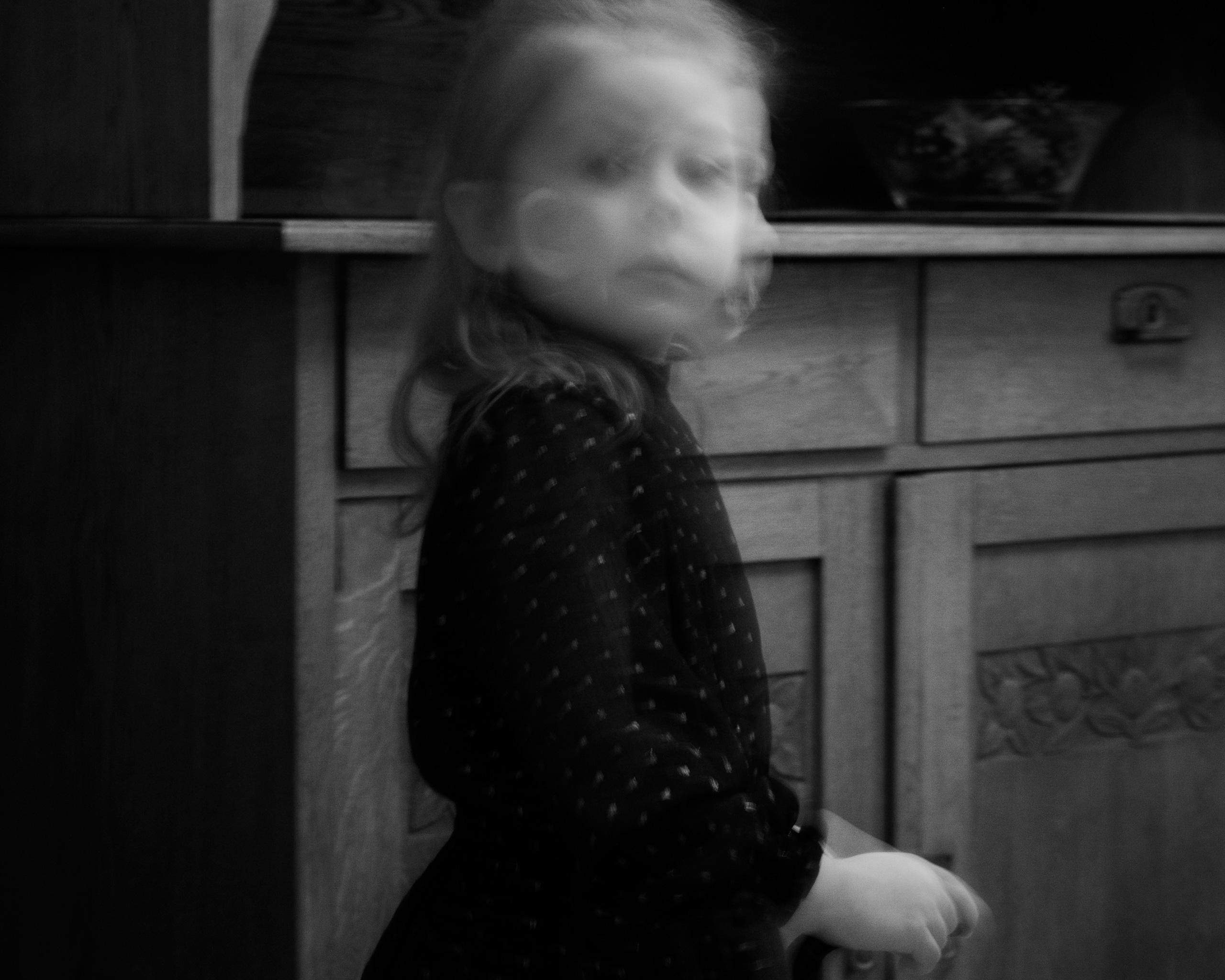 Black and white photo of a young girl in a dark patterned dress standing in front of a carved wooden cabinet, captured with motion blur that gives her face a ghostly, double-exposed appearance. The child’s expression appears contemplative or curious, with blurred facial features adding a surreal, haunting effect. Her long hair flows slightly with movement, and her hand is resting on an unseen object in the foreground.