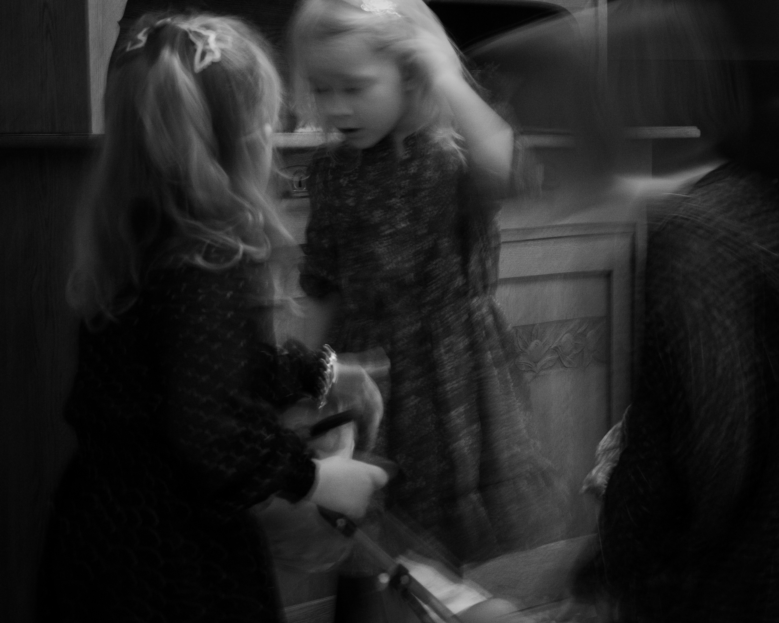 Black and white photo of a young girl with blonde hair in a patterned dress standing in front of a mirror, creating a ghostly double exposure effect; her reflection and blurred movements suggest motion or a dreamlike atmosphere. The child has a hair clip and appears to be brushing or touching her hair while looking at her reflection. The background features a wooden cabinet with carved floral detailing.