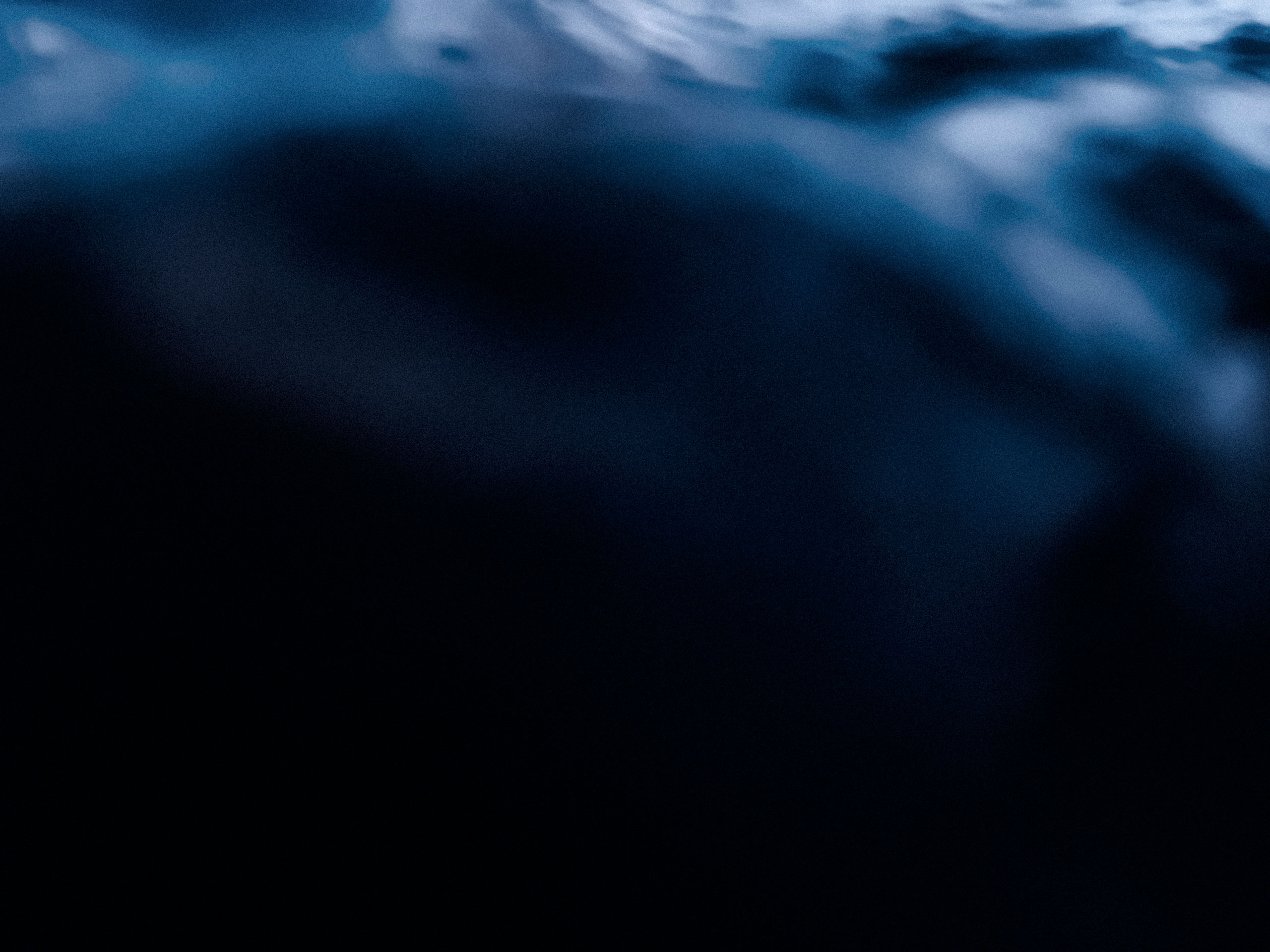 Abstract underwater photograph featuring a dark, shadowy ocean depth with soft gradients of blue and black, illuminated by diffused light from the surface above. The rippling water at the top creates a sense of motion and depth, enhancing the mysterious and immersive marine atmosphere.