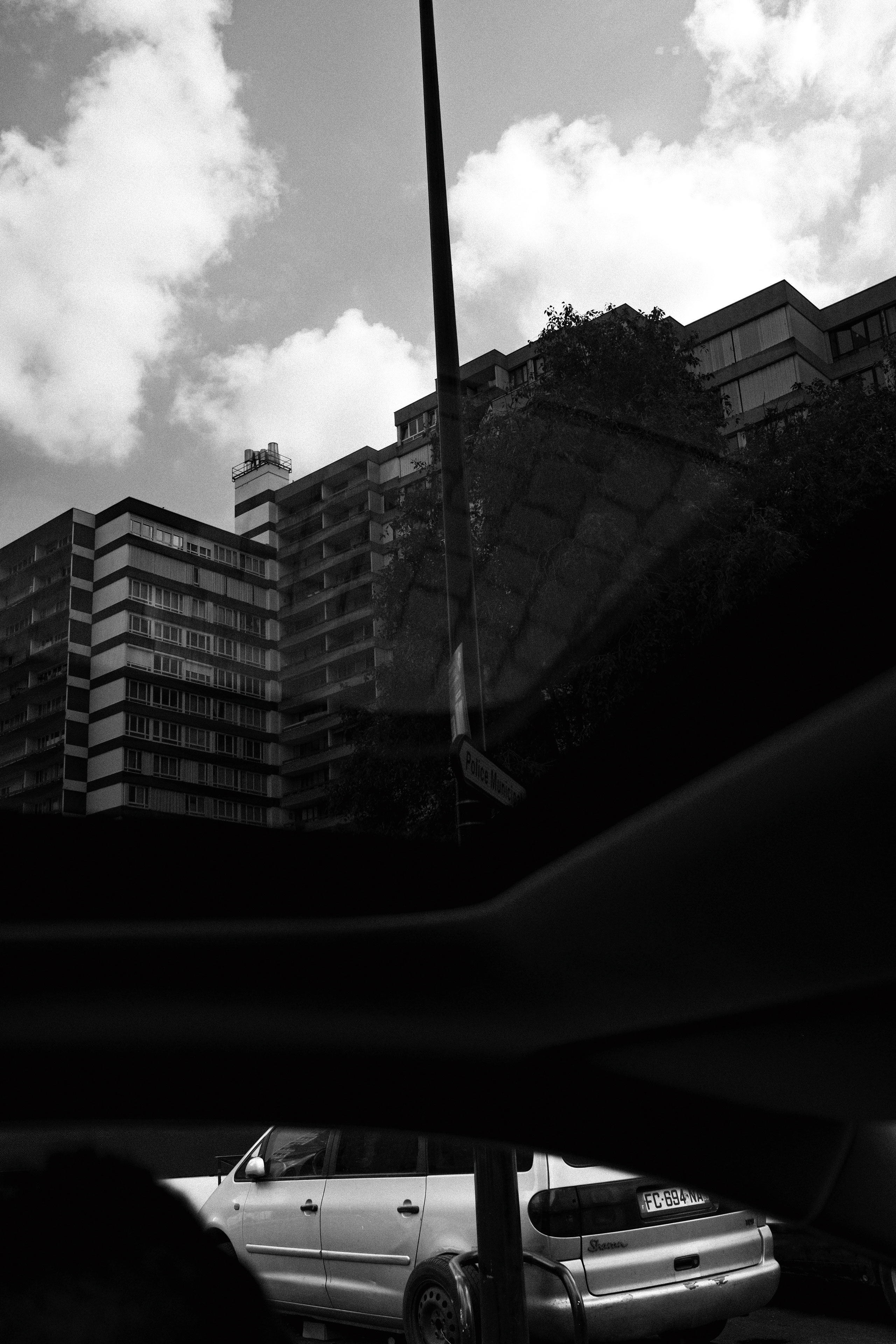Black and white street-level photograph capturing a perspective view of modern apartment buildings with geometric facades under a partly cloudy sky. A silver station wagon is parked in the foreground, partially obscured by the dark interior framing of a vehicle window. A street sign reading “Police Municipale” is visible near the center. The image features strong urban textures, reflections on glass, and a juxtaposition of architectural lines and organic cloud formations.