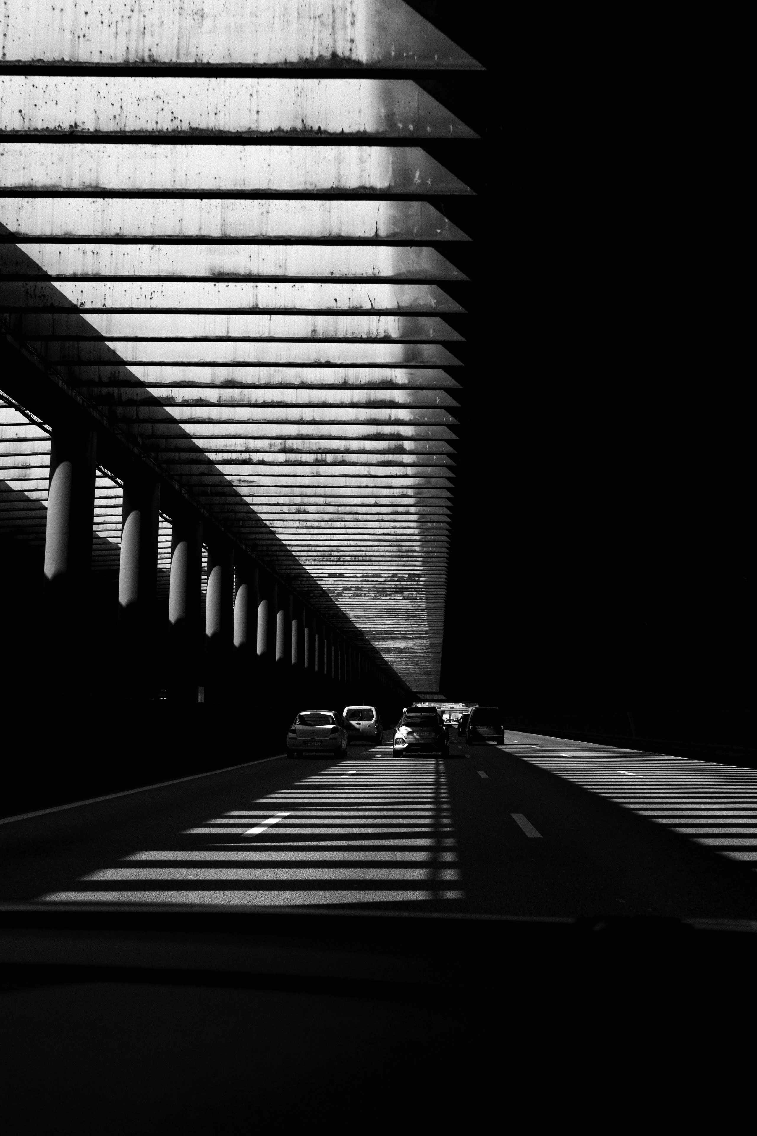 Black and white photo of a highway passing under a series of concrete beams, creating dramatic diagonal shadows across the road. Several vehicles are visible ahead, some parked with open doors, possibly indicating an incident or stop. The architectural elements and harsh contrast between light and shadow add a geometric and cinematic feel.