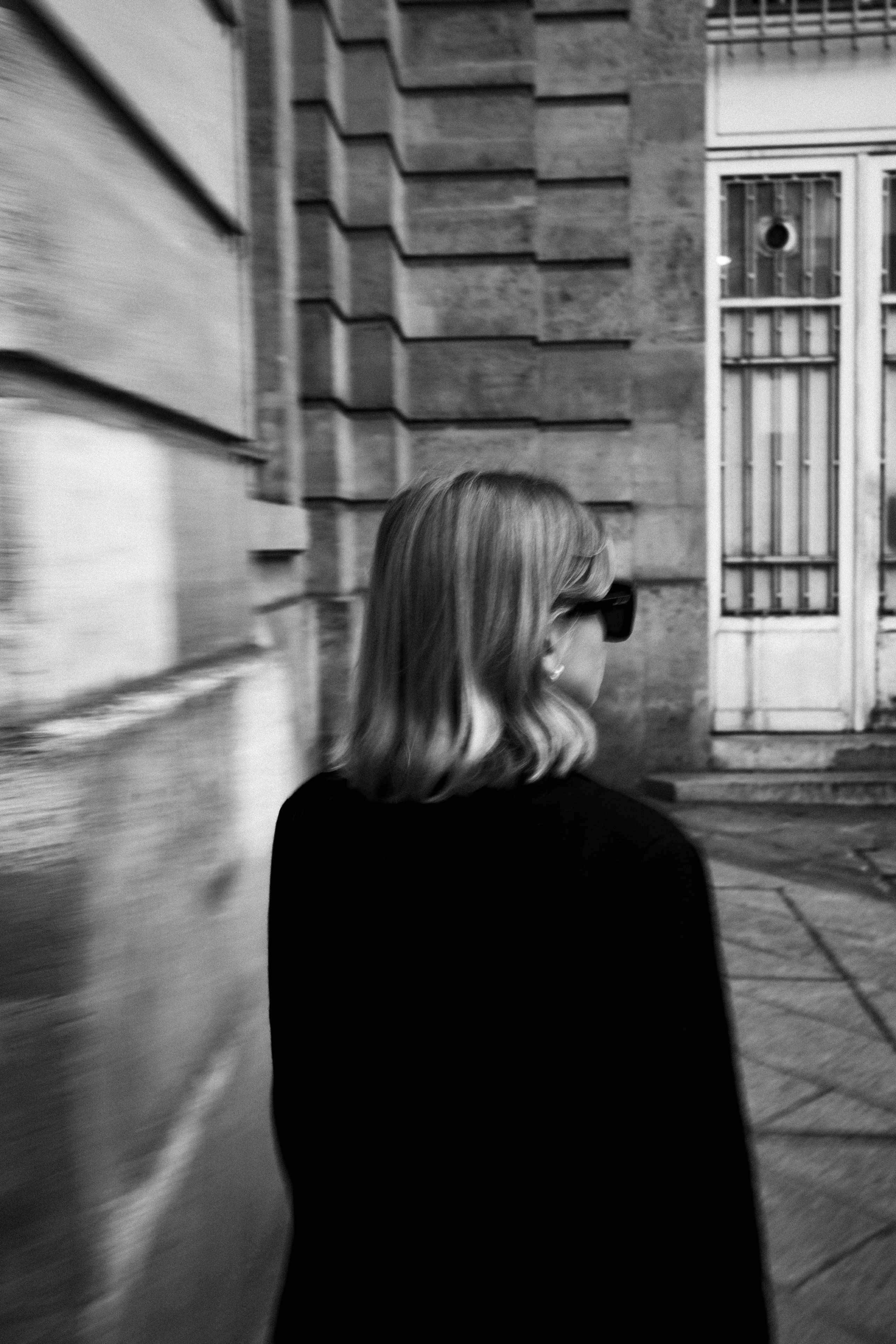 Black and white street photograph capturing the back view of a woman with shoulder-length blonde hair, wearing dark sunglasses and a black coat, walking alongside a historic stone building with a geometric, neoclassical facade. The image is slightly blurred, enhancing a sense of movement and anonymity. A barred white door and stone steps appear in the background, adding architectural detail.