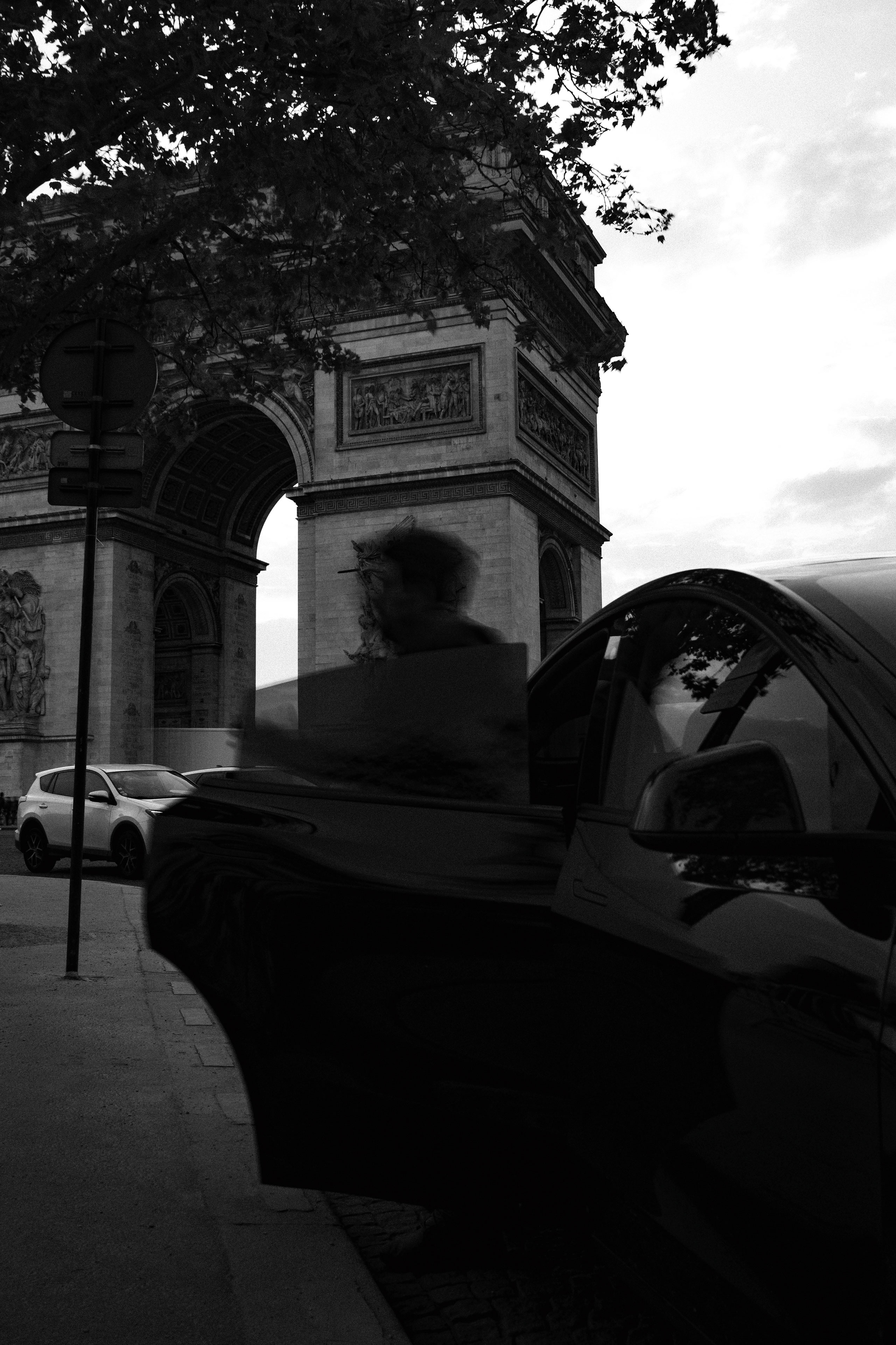 Black and white photo of the Arc de Triomphe in Paris captured at dusk, with a blurred figure stepping out of a sleek black car in the foreground. The monument’s neoclassical architecture, sculptural reliefs, and intricate details are partially shaded by overhanging tree branches. The contrast between the historical arch and the modern vehicle emphasizes the blend of old and new in urban Paris.