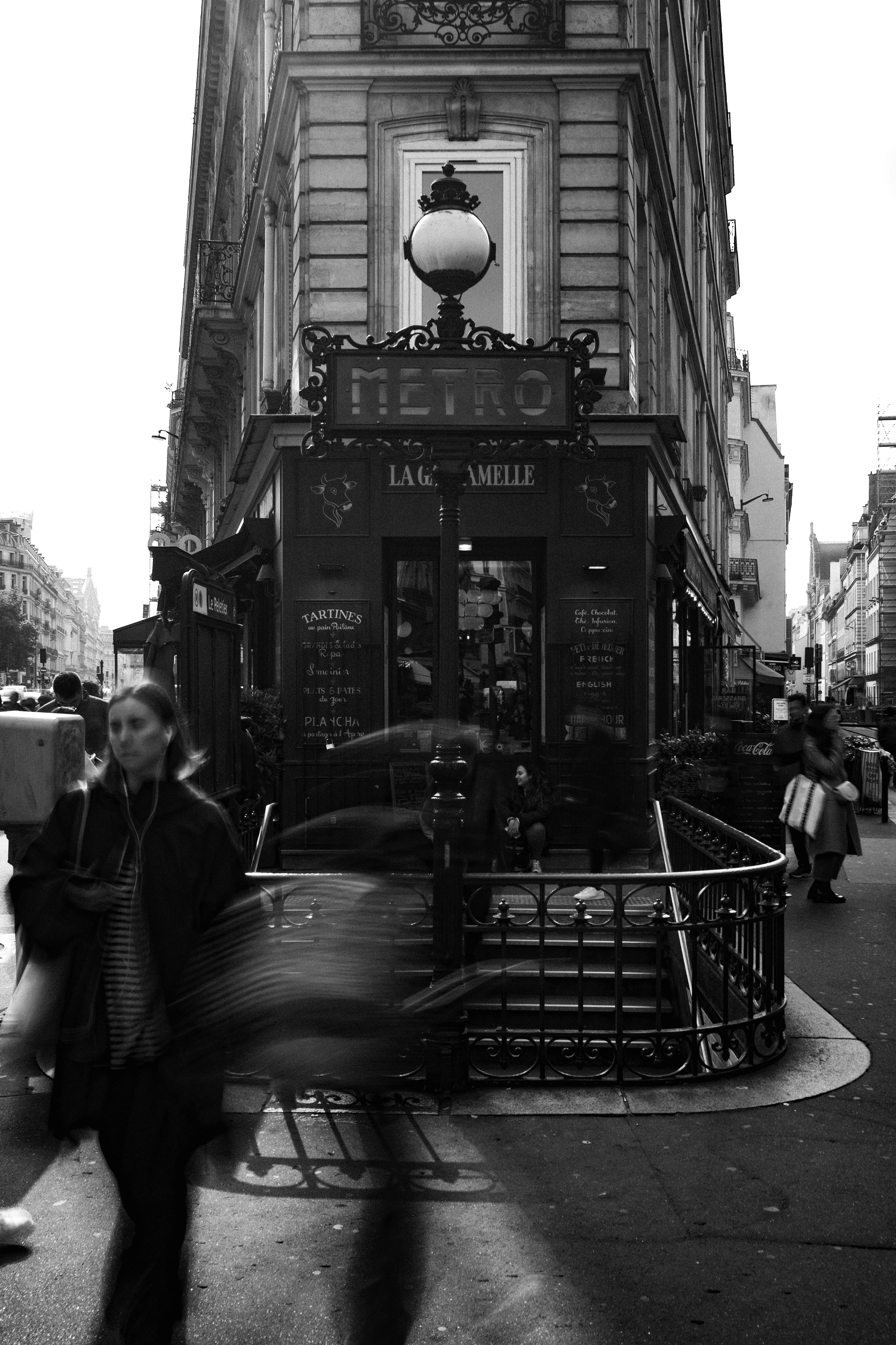 Black and white photograph capturing a bustling Parisian street scene in front of a classic Métro entrance. The ornate iron railing and vintage lamp post with a “METRO” sign mark the entrance, positioned in front of a building housing the restaurant “La Gamelle.” Blurred motion from pedestrians walking past contrasts with a stationary woman seated on the steps. The backdrop includes Haussmann-style architecture and street signs indicating the “Opéra” metro station.
