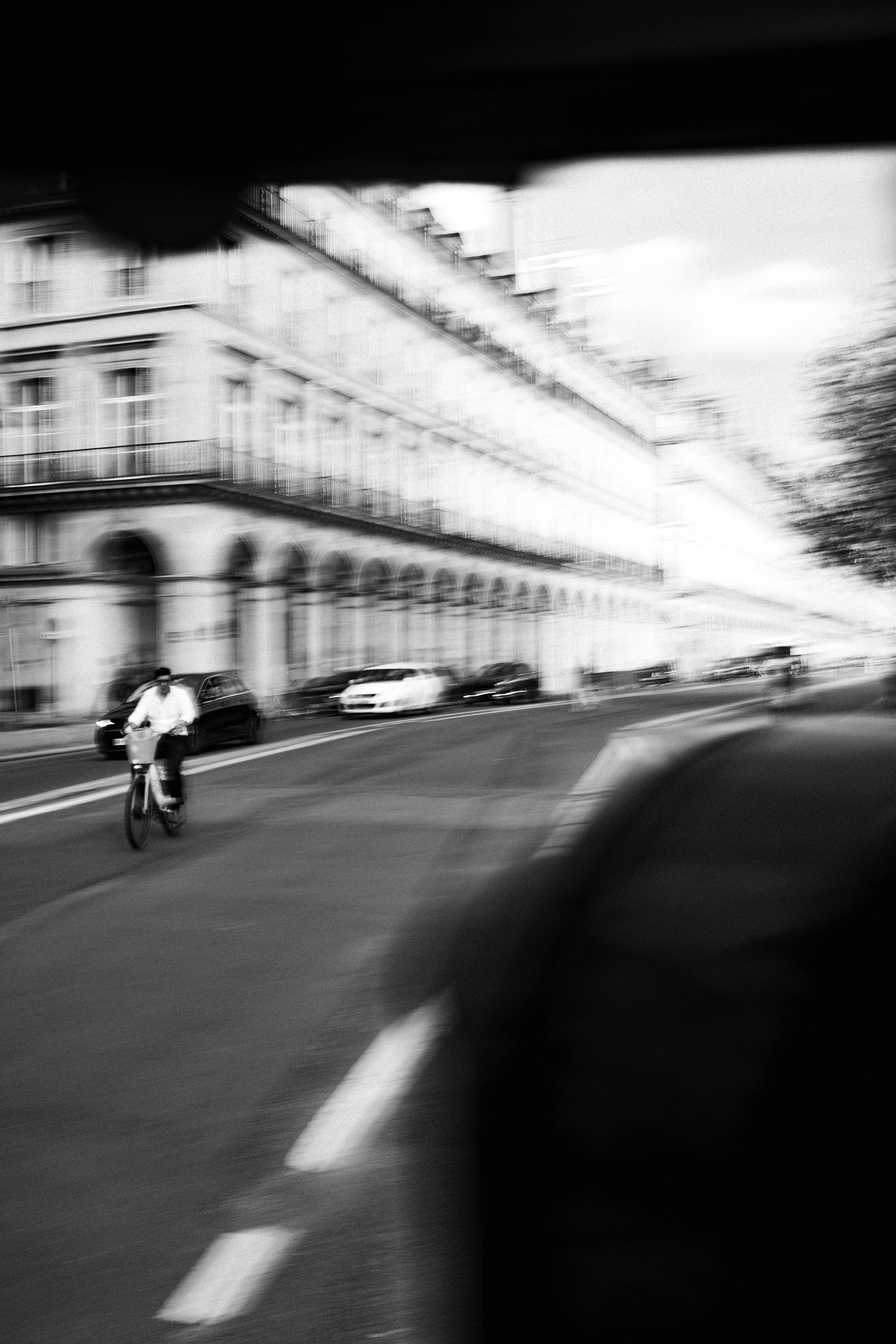 Black and white photo capturing motion blur of a cyclist riding along a Parisian street lined with neoclassical buildings featuring arched arcades and wrought iron balconies. The perspective emphasizes the vanishing point of the long, symmetrical façade. Cars are parked along the curb, and shadows from an overhead structure frame the image.