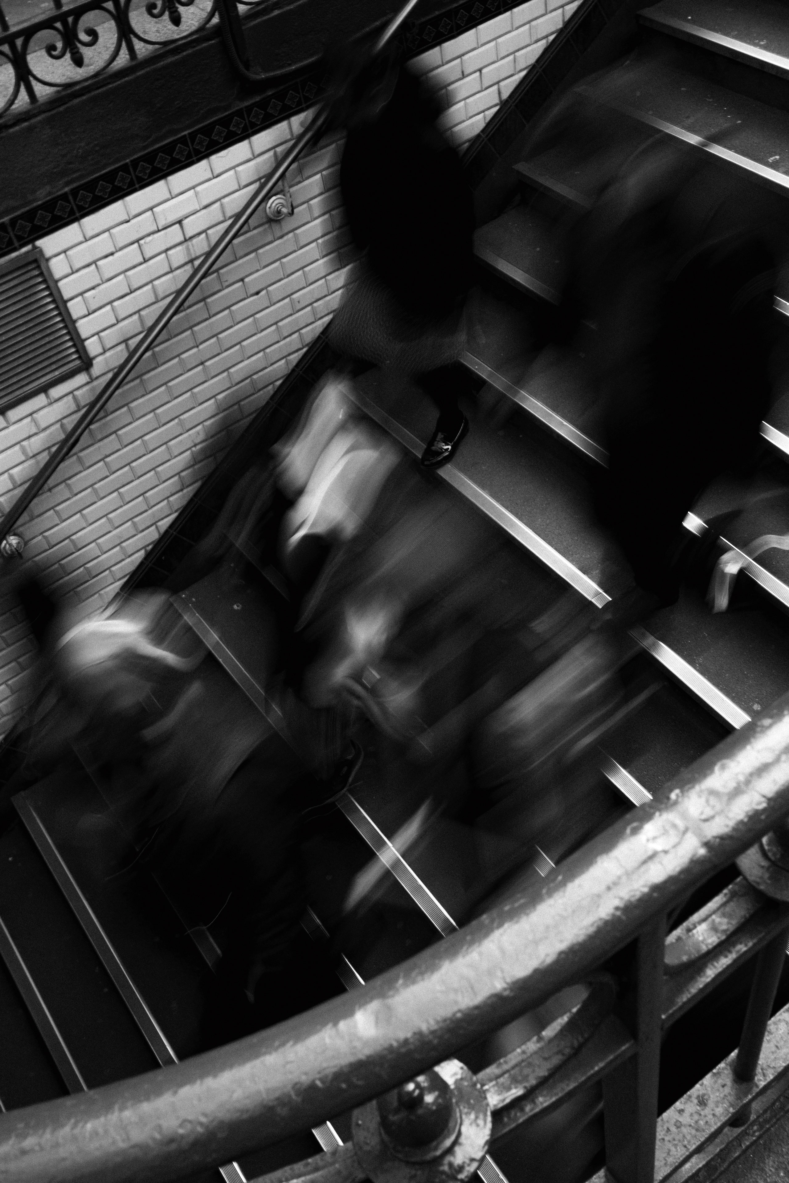 Black and white photograph capturing blurred motion of commuters walking up and down tiled stairs in a metro station. The image conveys dynamic energy and urban rhythm through long exposure, with streaked silhouettes and indistinct forms of people in constant movement. The stairs are bordered by classic white subway tiles with a decorative black trim and an ornate wrought iron railing, characteristic of Paris Métro stations.