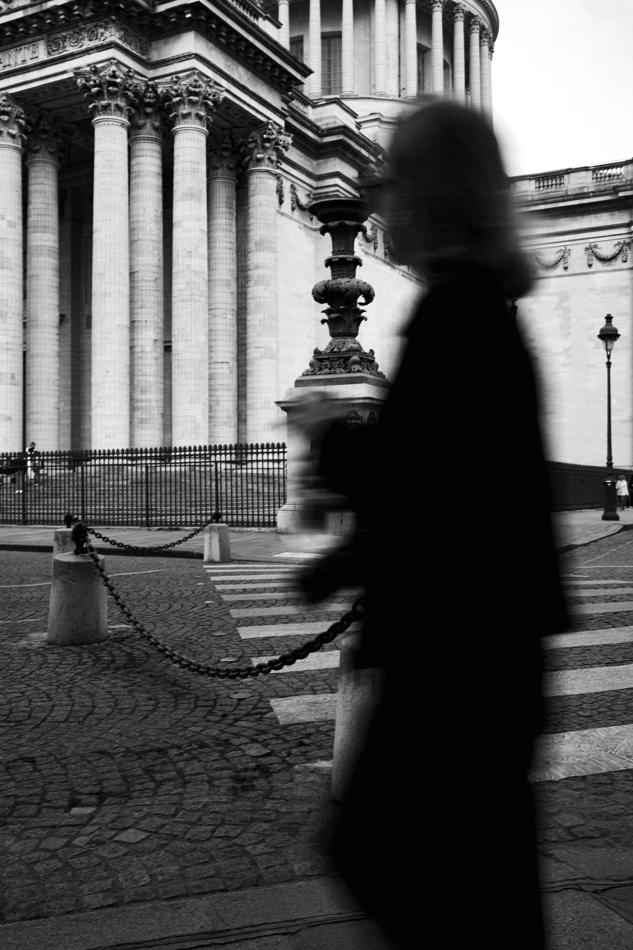 Black and white image of a neoclassical building, likely the Panthéon in Paris, with grand Corinthian columns and ornate architectural details. In the foreground, a blurred silhouette of a woman in motion dominates the right side, adding contrast and dynamic movement against the sharp, monumental backdrop. Cobblestone pavement, a pedestrian crosswalk, and chain-linked posts enhance the urban texture.