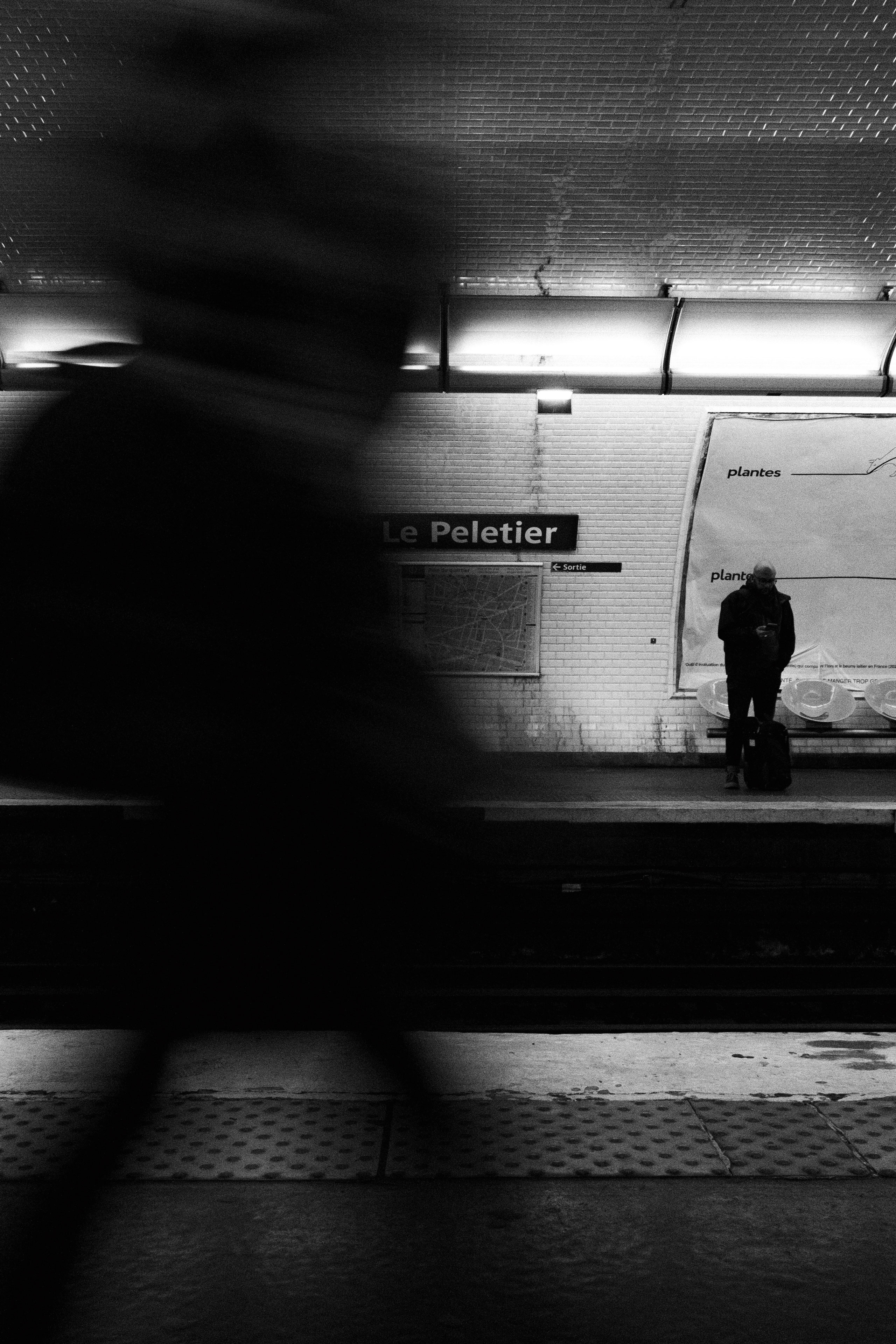 Black and white photo of Le Peletier Paris Métro station platform. A man stands still on the opposite side, looking at his phone with a suitcase beside him, while a blurry pedestrian in motion passes in the foreground. The platform is lined with white tiles, partially covered posters, and illuminated by fluorescent ceiling lights. The motion blur adds a sense of urban urgency and contrasts with the lone still figure.