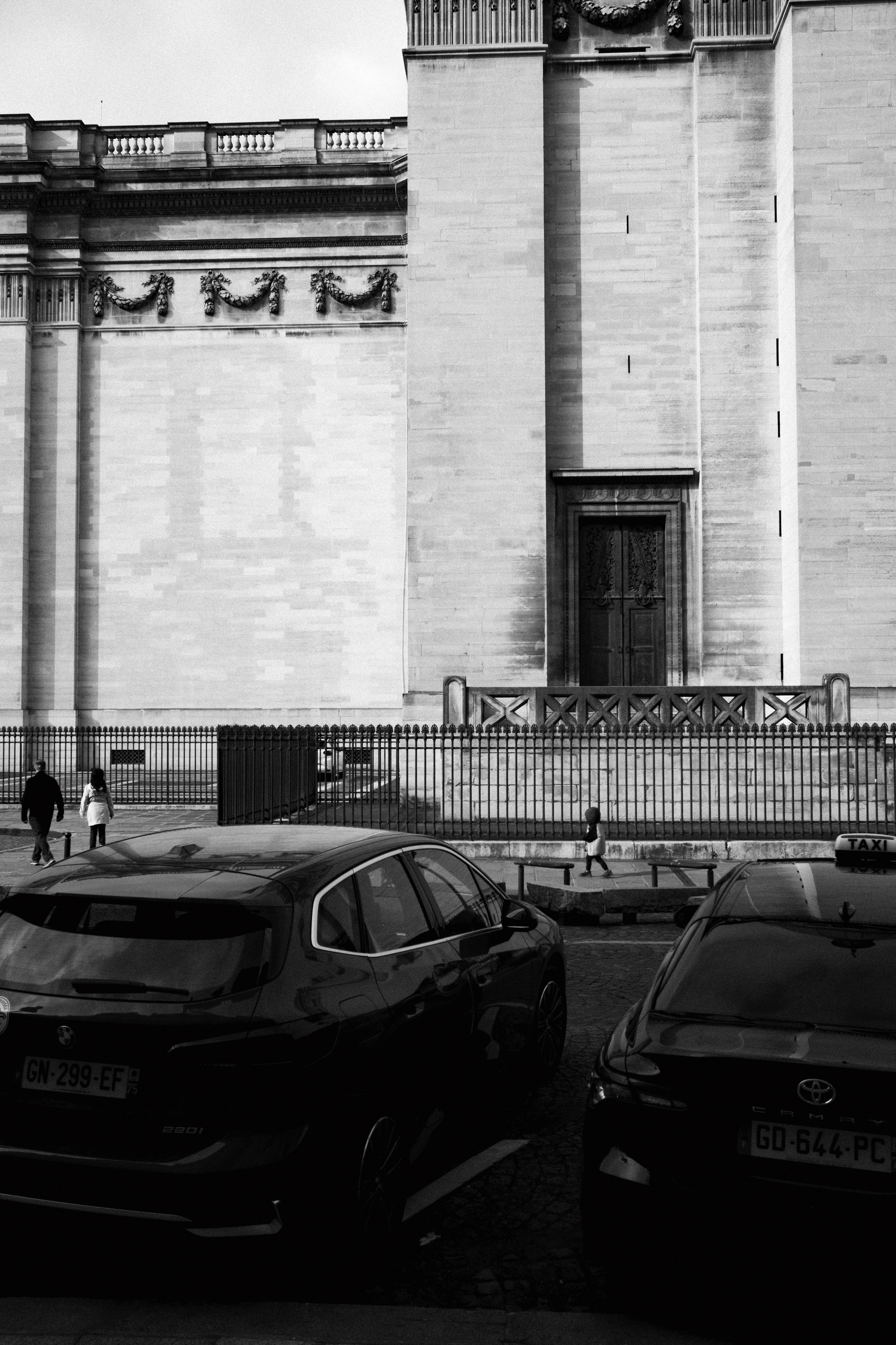 Black and white street photo showing the exterior of a grand neoclassical building with tall columns, ornate stone carvings, and a large wooden door at its center. In the foreground, two dark-colored parked cars, one a BMW and the other a Toyota taxi, frame the bottom edge of the image. Several pedestrians, including a child with a balloon, walk along the sidewalk behind a wrought iron fence.
