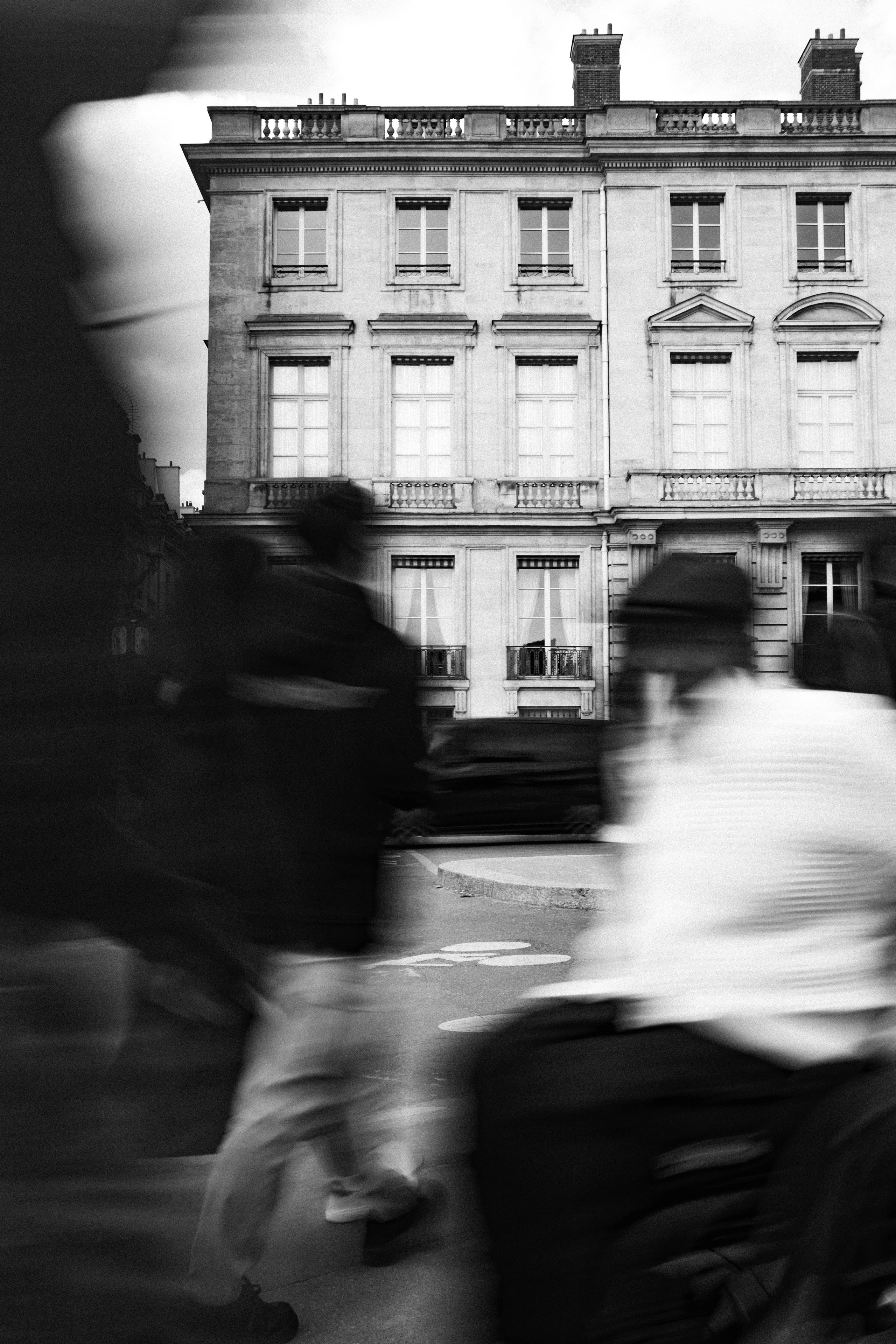 Black and white street photo showing blurred pedestrians walking in front of a stately Parisian building with classic architectural elements like tall windows, wrought iron balconies, and decorative stonework. The people in motion, including a figure in a wheelchair or stroller, create a dynamic foreground against the stationary elegance of the historic façade.