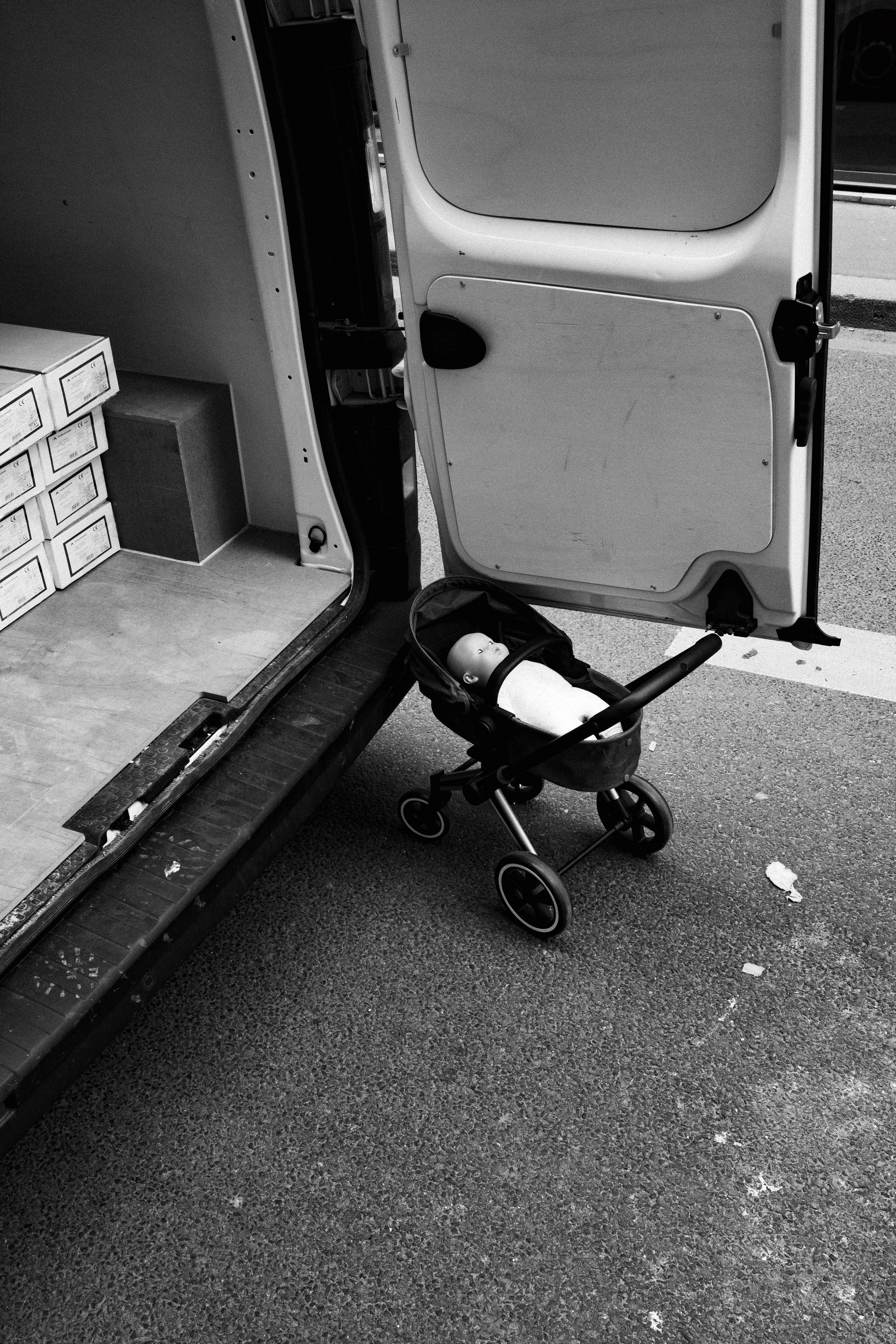 Black and white photo depicting an open delivery van parked on a street, with a baby doll placed in a small stroller positioned behind the open rear door. Inside the van, neatly stacked boxes are visible, suggesting a commercial or logistic setting. The doll’s placement in the stroller adds an unsettling or surreal touch to an otherwise ordinary urban scene.