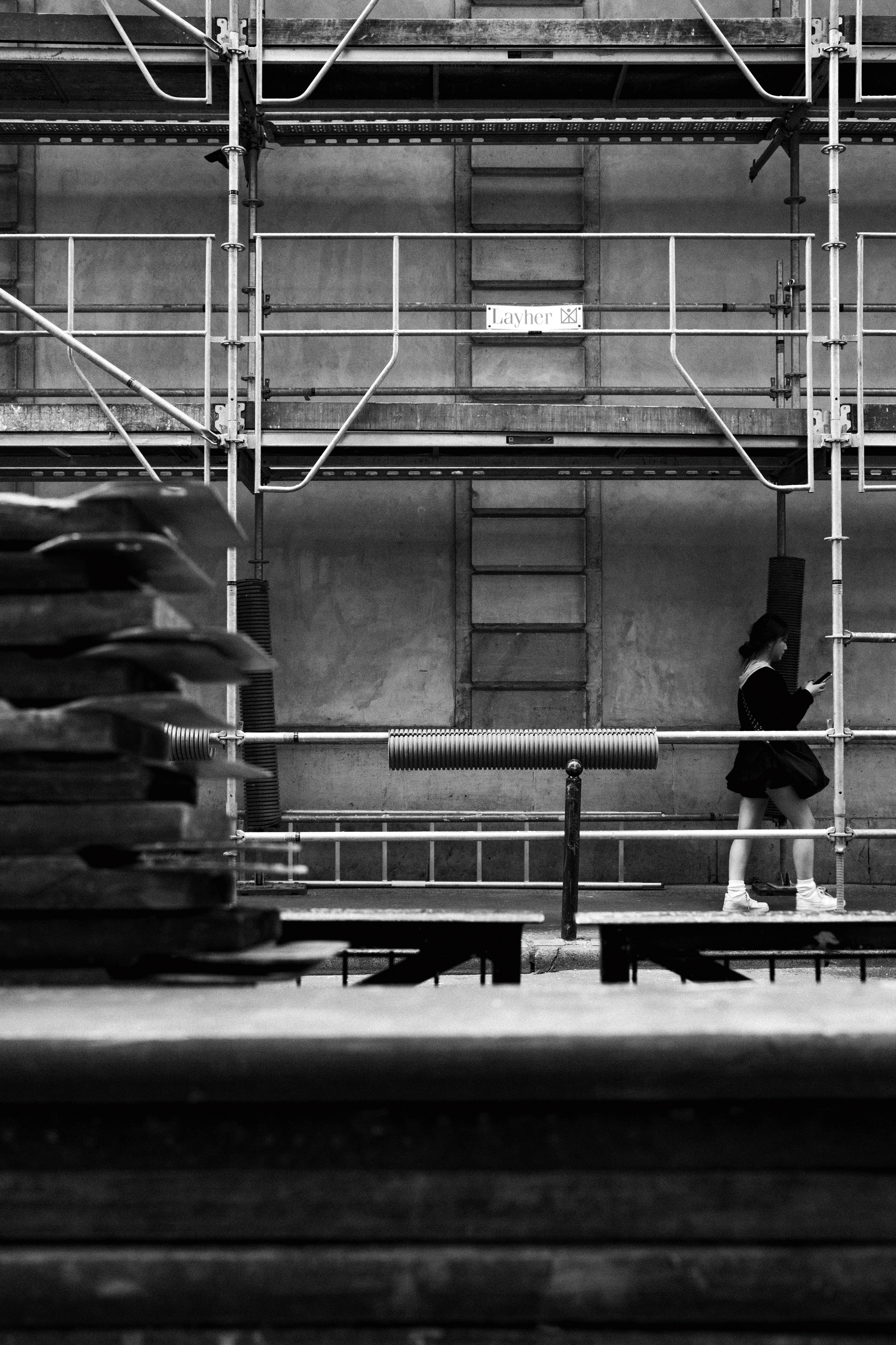 Black and white urban photo of a woman walking past a construction site, framed by metal scaffolding with the brand name “Layher” visible on a crossbar. The woman, dressed in a skirt and sneakers, appears to be looking at her phone while passing through the grid-like structure. In the foreground, stacks of wooden planks and construction materials add texture and depth. The contrast between the rigid industrial framework and the motion of the pedestrian creates a visually dynamic street scene.