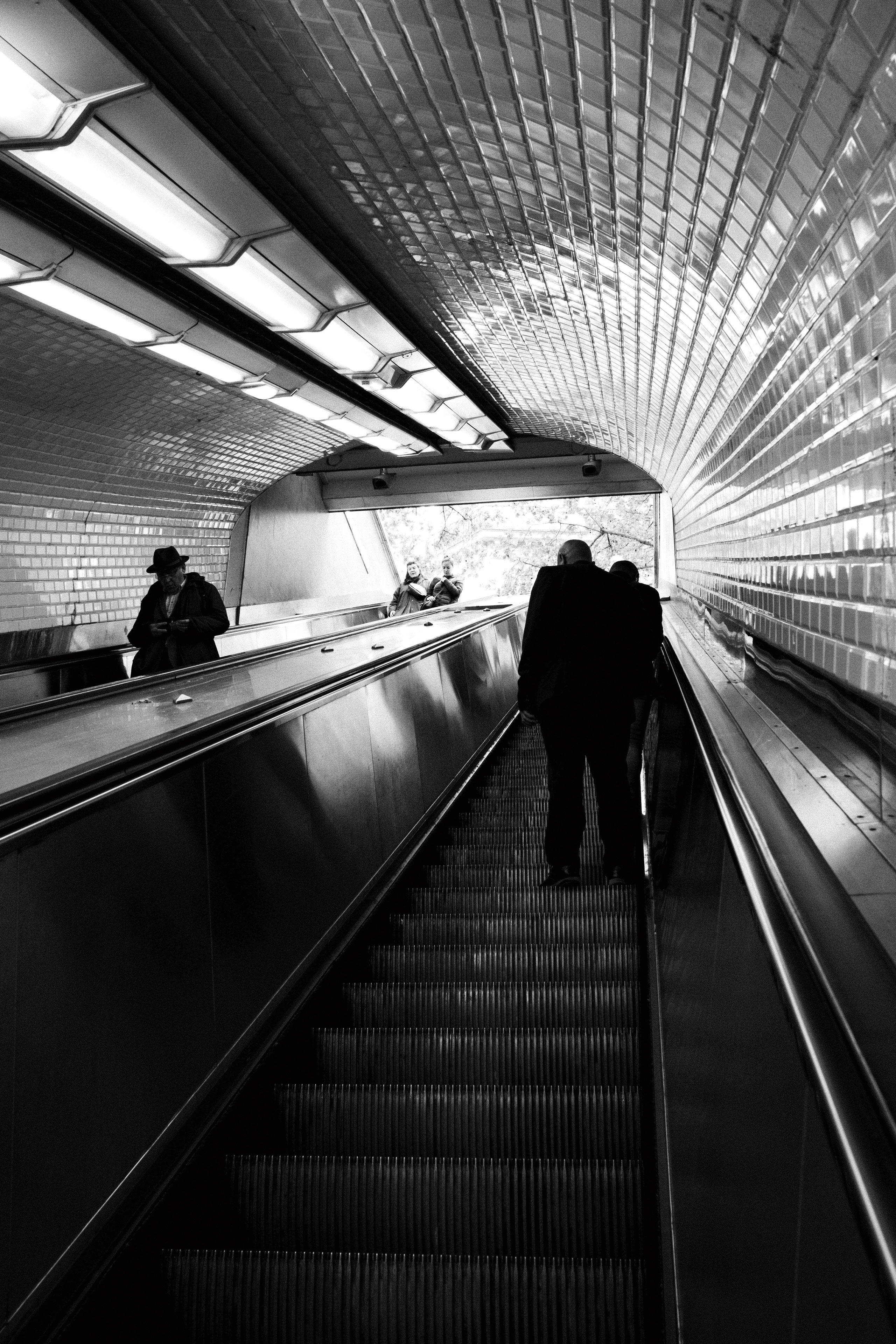 Black and white photograph of an upward-moving escalator in a Paris Métro station, with reflective tiled walls and ceiling curving around the tunnel. Several people are seen ascending—some on the escalator and others walking beside it on the ramp—creating a layered sense of movement and depth. The backlit station exit reveals a glimpse of trees outside, contrasting the dim interior with natural light.