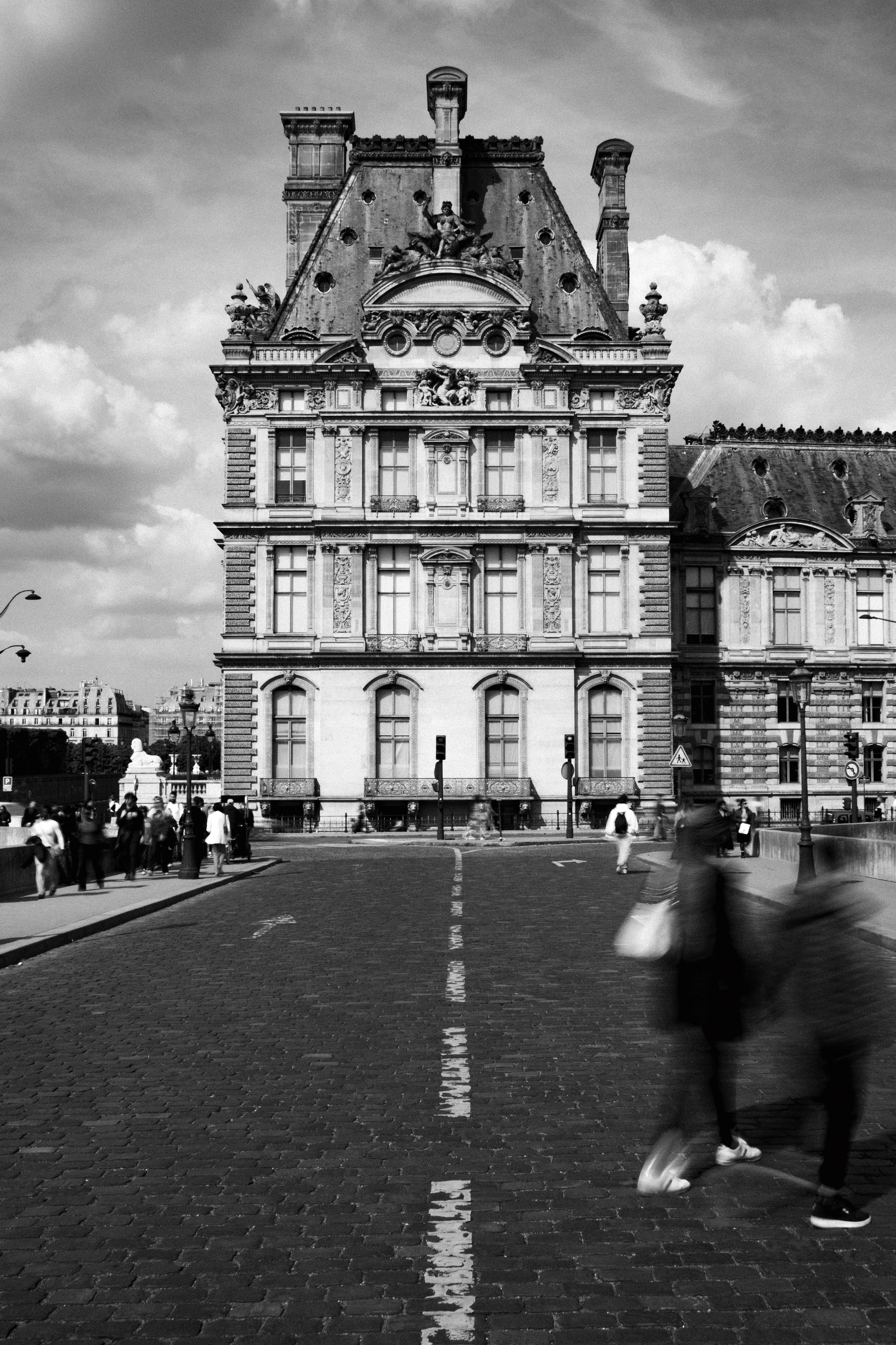 Black and white photograph of the richly ornamented Pavillon de Flore, part of the Louvre Palace in Paris, viewed from a cobblestone bridge. The symmetrical Renaissance Revival facade features elaborate sculptures, arched windows, and decorative chimneys. A group of pedestrians, some blurred in motion, cross the foreground, contrasting the still grandeur of the architecture.