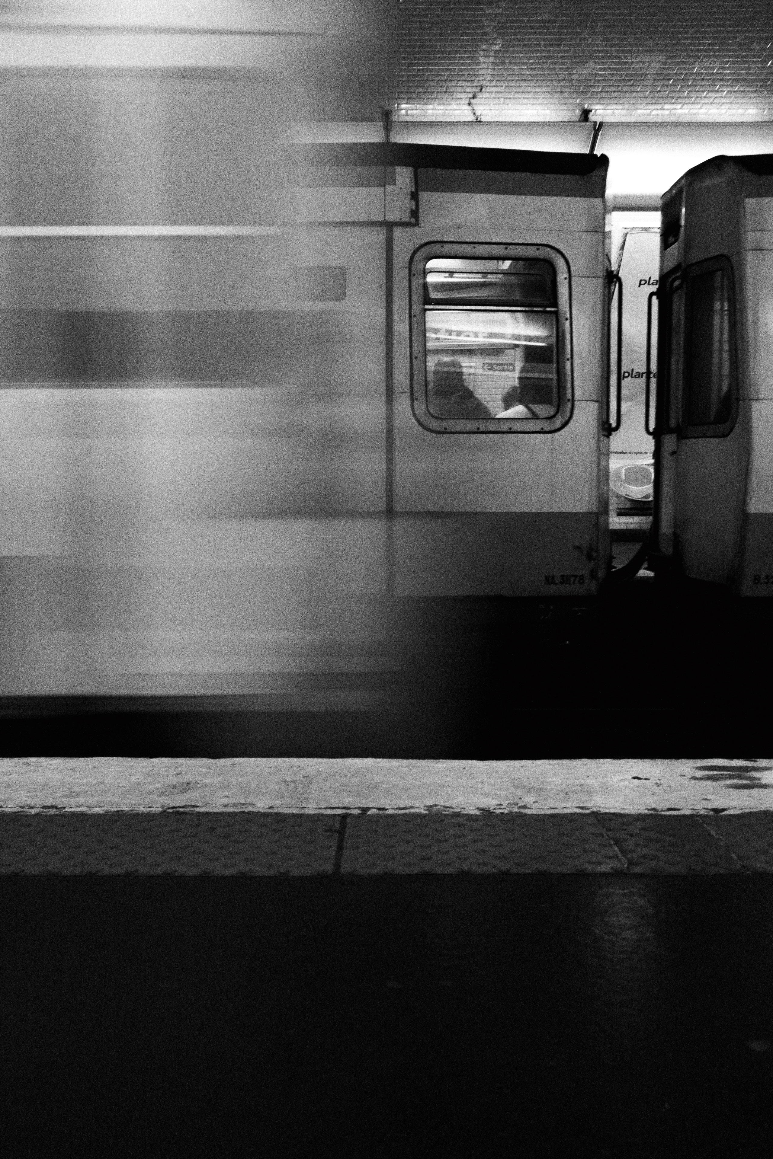 Black and white photo of a Paris Métro platform capturing two stationary train cars with a third in motion passing in front. The blurred streak of the moving train dominates the left side, creating a dynamic contrast with the stillness of the car on the right, where silhouettes of seated passengers are visible through the window. The tiled station wall and dim overhead lighting add texture and atmosphere.
