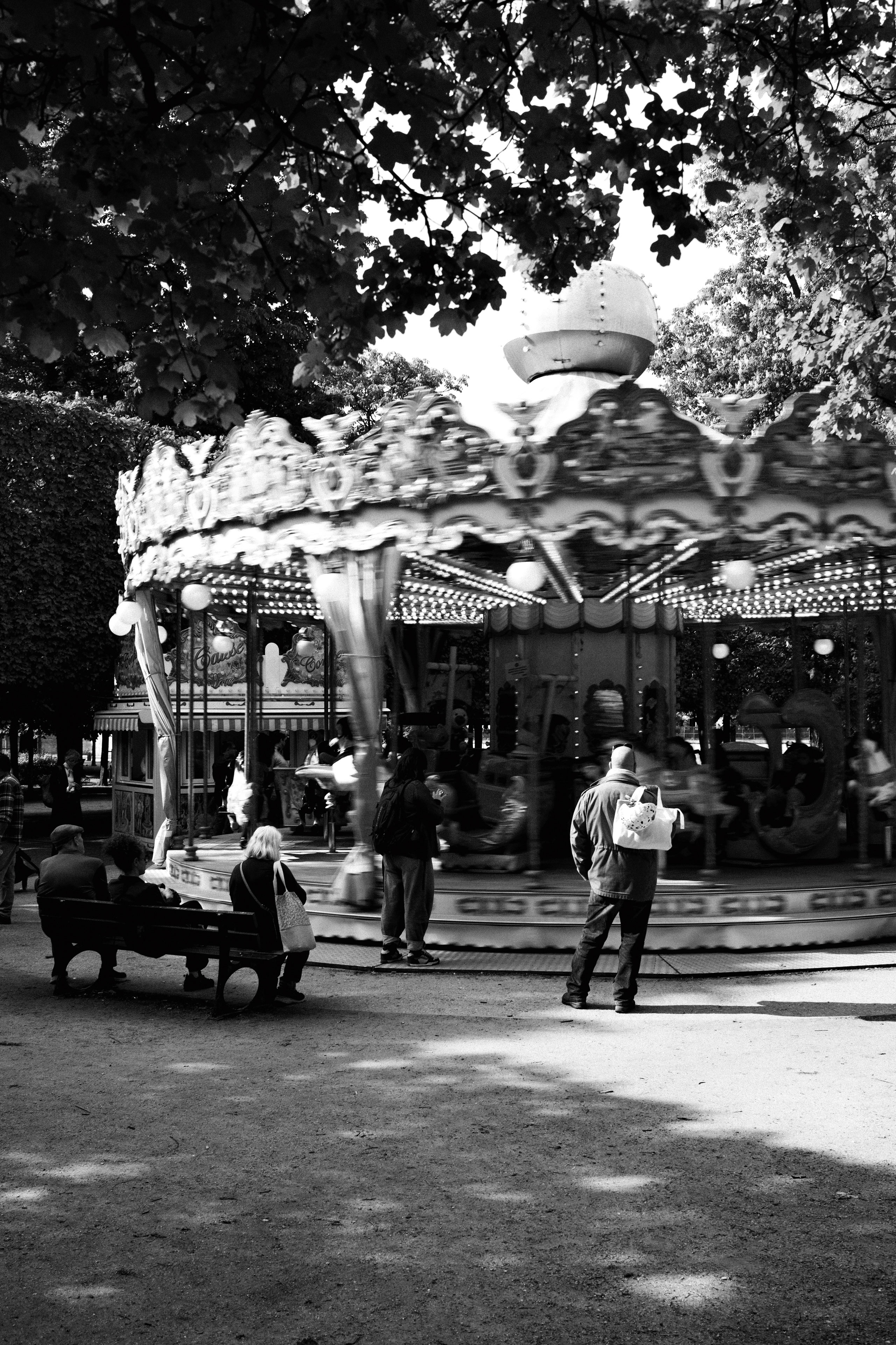 Black and white photo of a vintage carousel in motion at a shaded park, likely in Paris. The ornate merry-go-round is adorned with decorative horses and illuminated string lights, spinning with children aboard. Adults stand nearby, watching, while a few people sit on benches under the tree canopy. The blurred motion of the ride contrasts with the stillness of the bystanders.