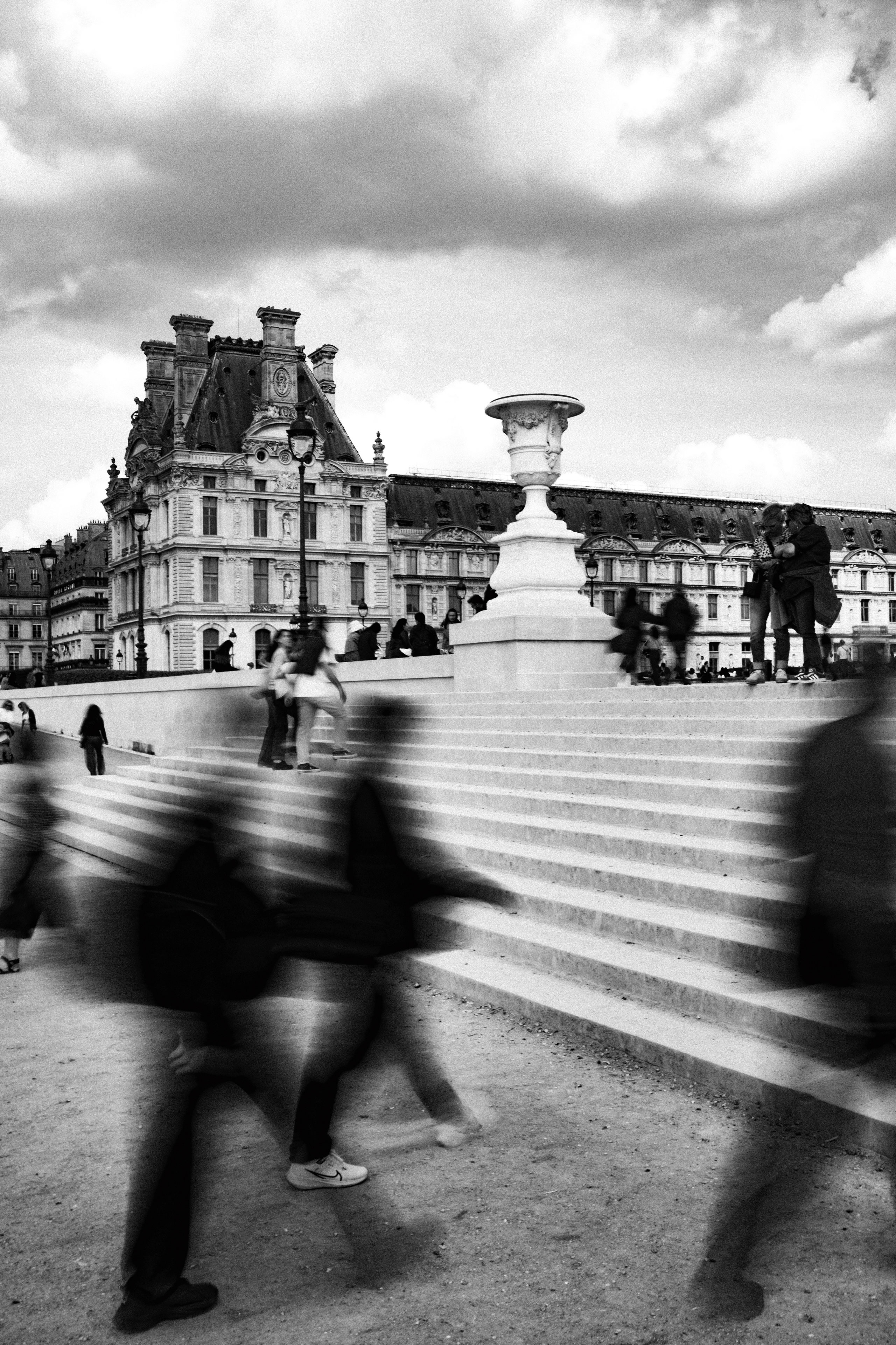 Black and white photo of a busy staircase at the Jardin des Tuileries in Paris, with people blurred in motion in the foreground and others gathered atop the stairs. A large classical urn stands centered above the steps, with the ornate Renaissance architecture of the Louvre Palace visible in the background. The image captures a juxtaposition of historical grandeur and modern life, with dynamic human movement set against still, majestic stonework.