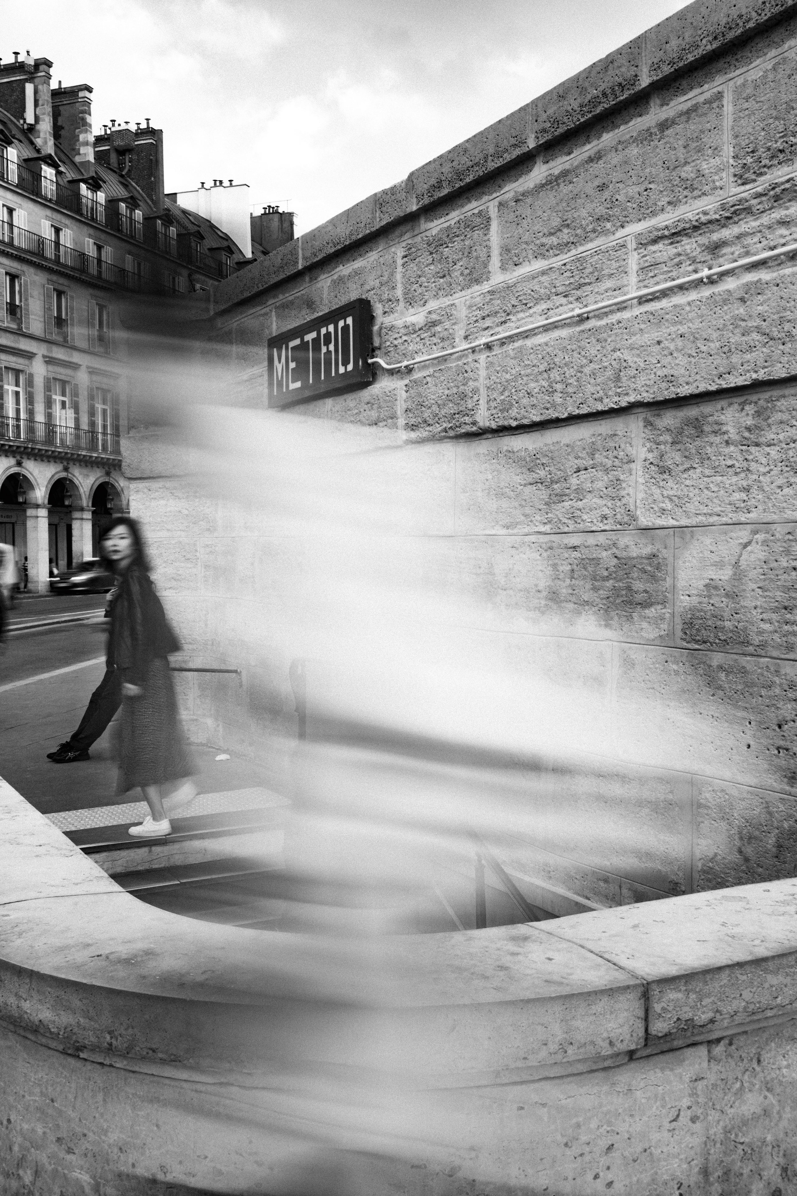 Black and white photo capturing a Paris Métro entrance with a woman pausing at the top of the stairs. A motion blur of passing pedestrians creates ghostly streaks of movement across the foreground, contrasting with the static elements of stone architecture and a vintage “METRO” sign mounted on the textured wall. The background reveals classic Parisian buildings with wrought iron balconies and arched windows.