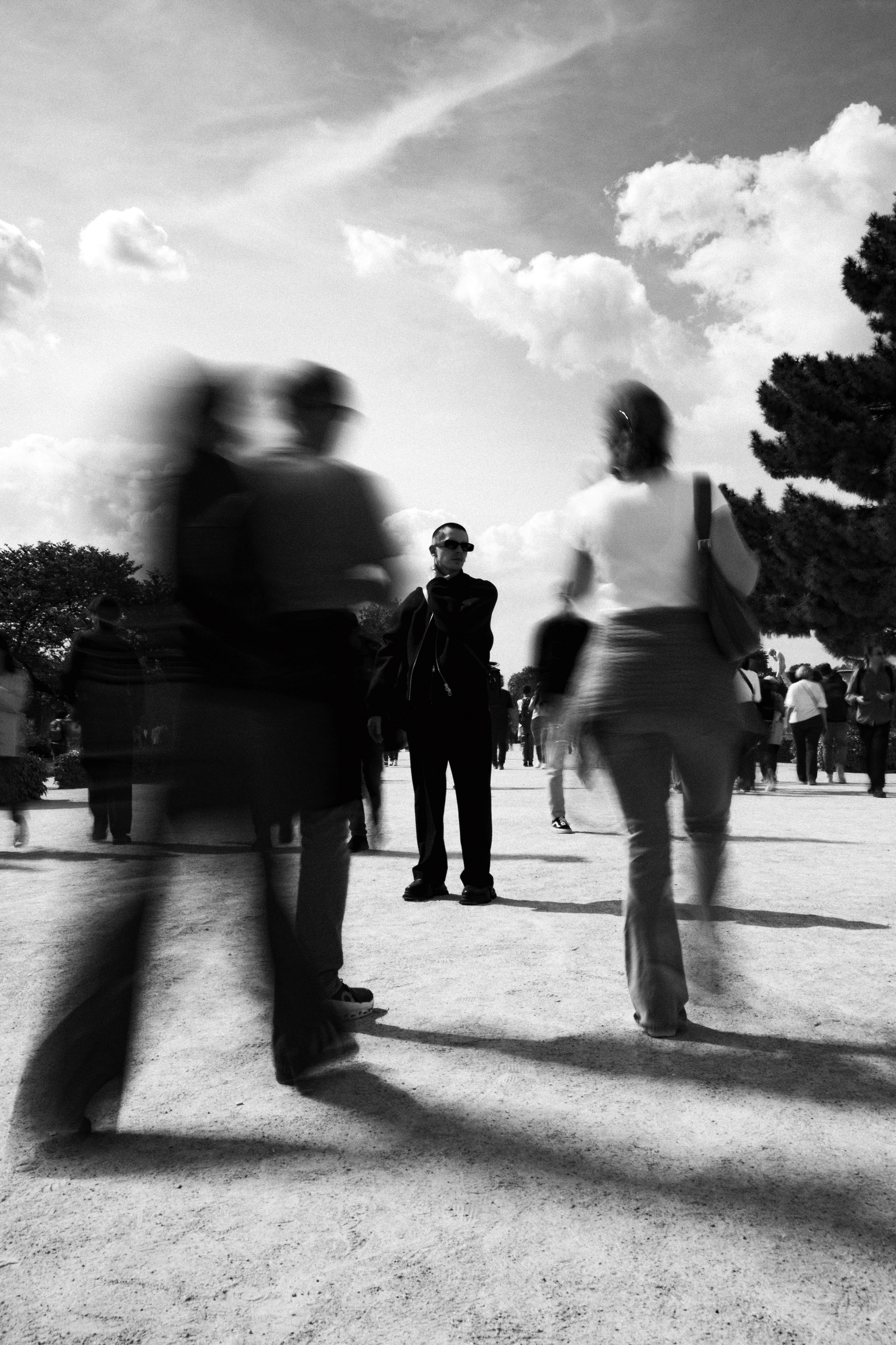 Black and white photograph capturing a solitary man in dark clothing and sunglasses standing still in the Jardin des Tuileries, Paris, surrounded by a crowd in motion blur. The contrast between his stillness and the blurred figures walking past him creates a striking sense of calm amidst chaos. The bright sky with scattered clouds and soft shadows on the ground enhance the dramatic, contemplative atmosphere.