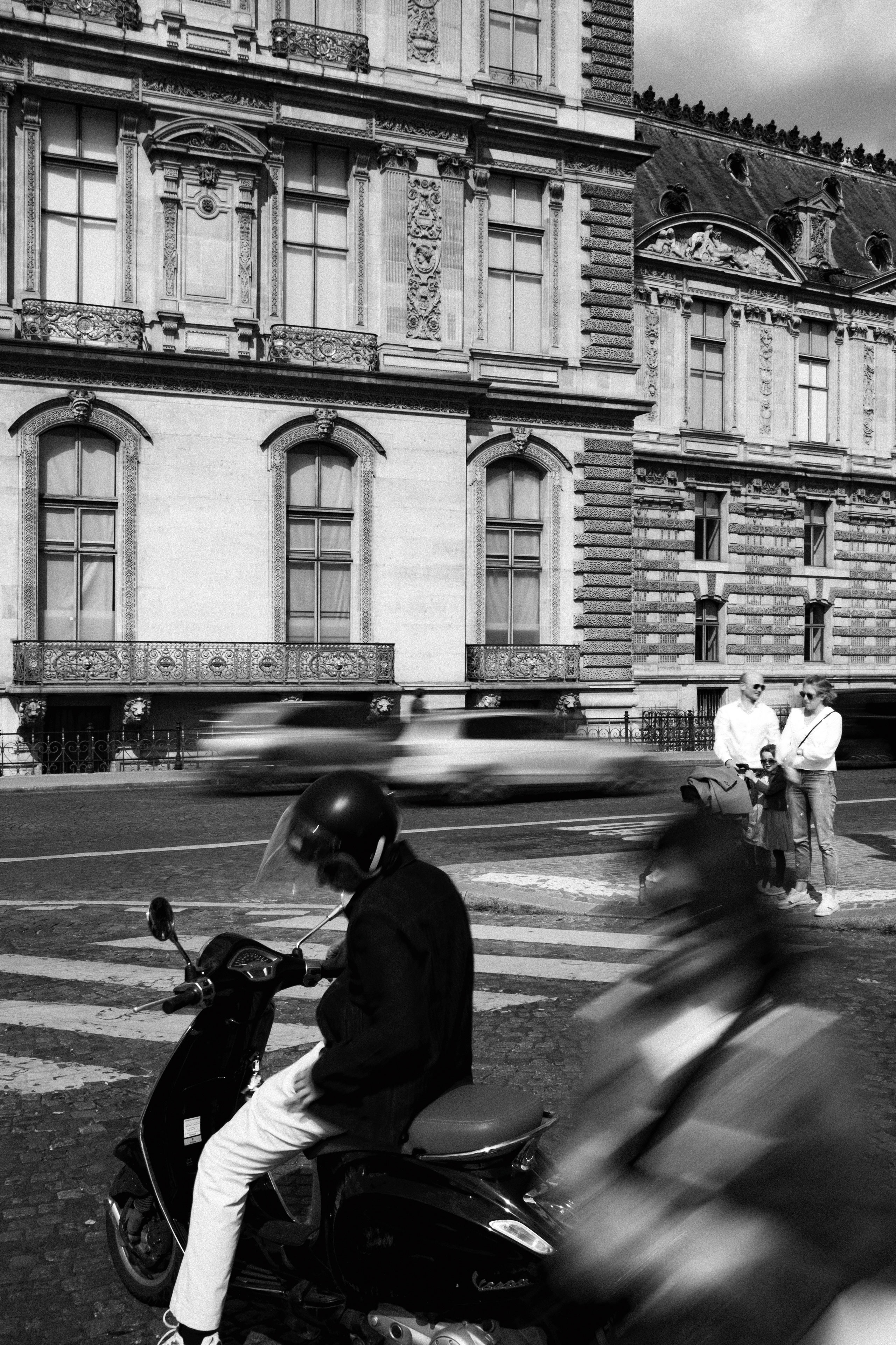 Black and white urban scene depicting motion and stillness in front of the ornate façade of the Louvre Palace in Paris. A man sits on a Vespa scooter in the foreground, while another scooter rider blurs past, capturing movement. Cars streak across the frame in the background, while a stationary family with a stroller watches from the sidewalk. The detailed Renaissance-style architecture serves as a grand backdrop.