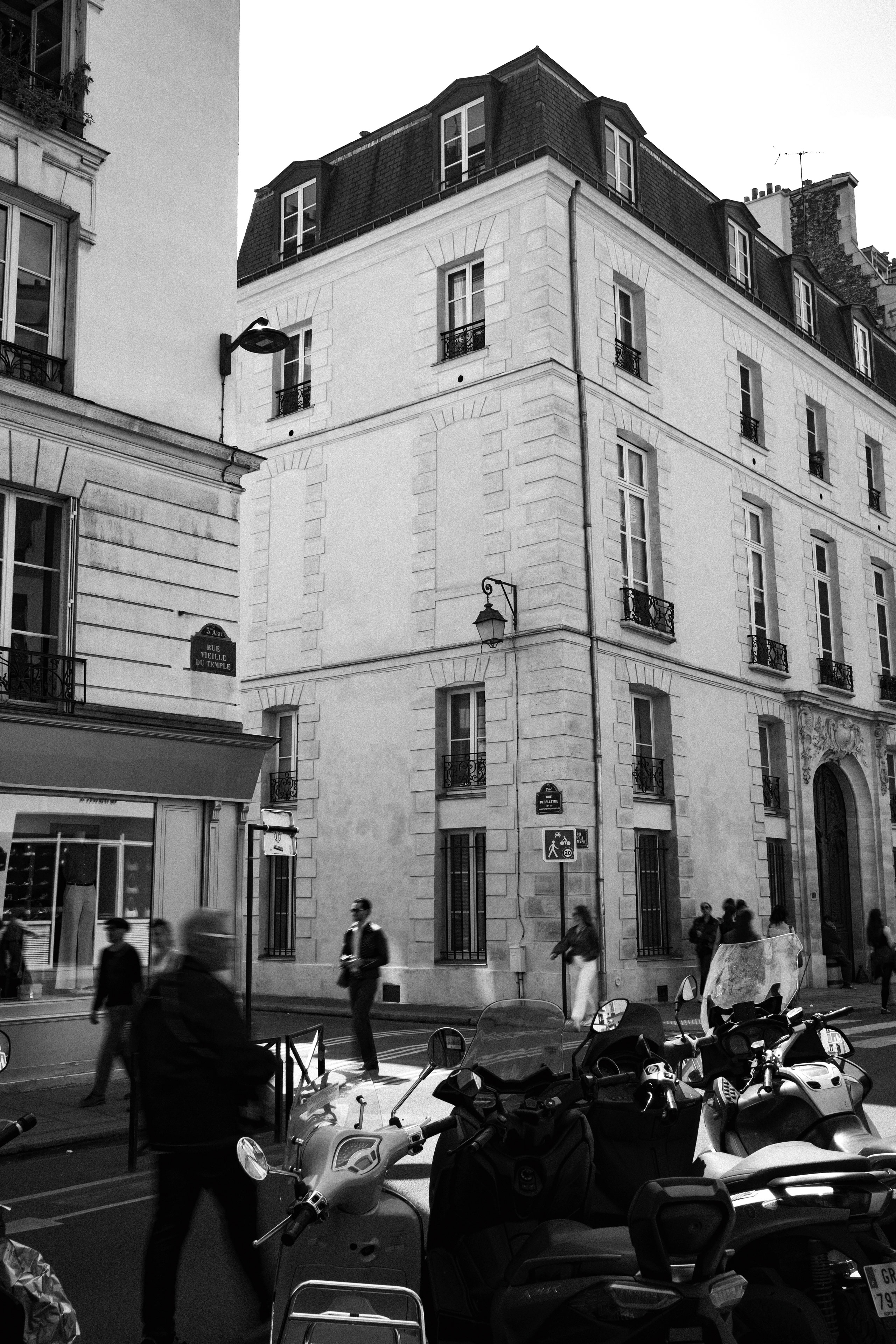 Black and white street scene capturing a corner in Paris with classic Haussmann-style architecture. The image features a prominent building at the intersection of Rue Vieille du Temple, with traditional French windows and wrought iron balconies. In the foreground, scooters and mopeds are parked along the street, while blurred pedestrians walk by, adding a sense of motion and city life.