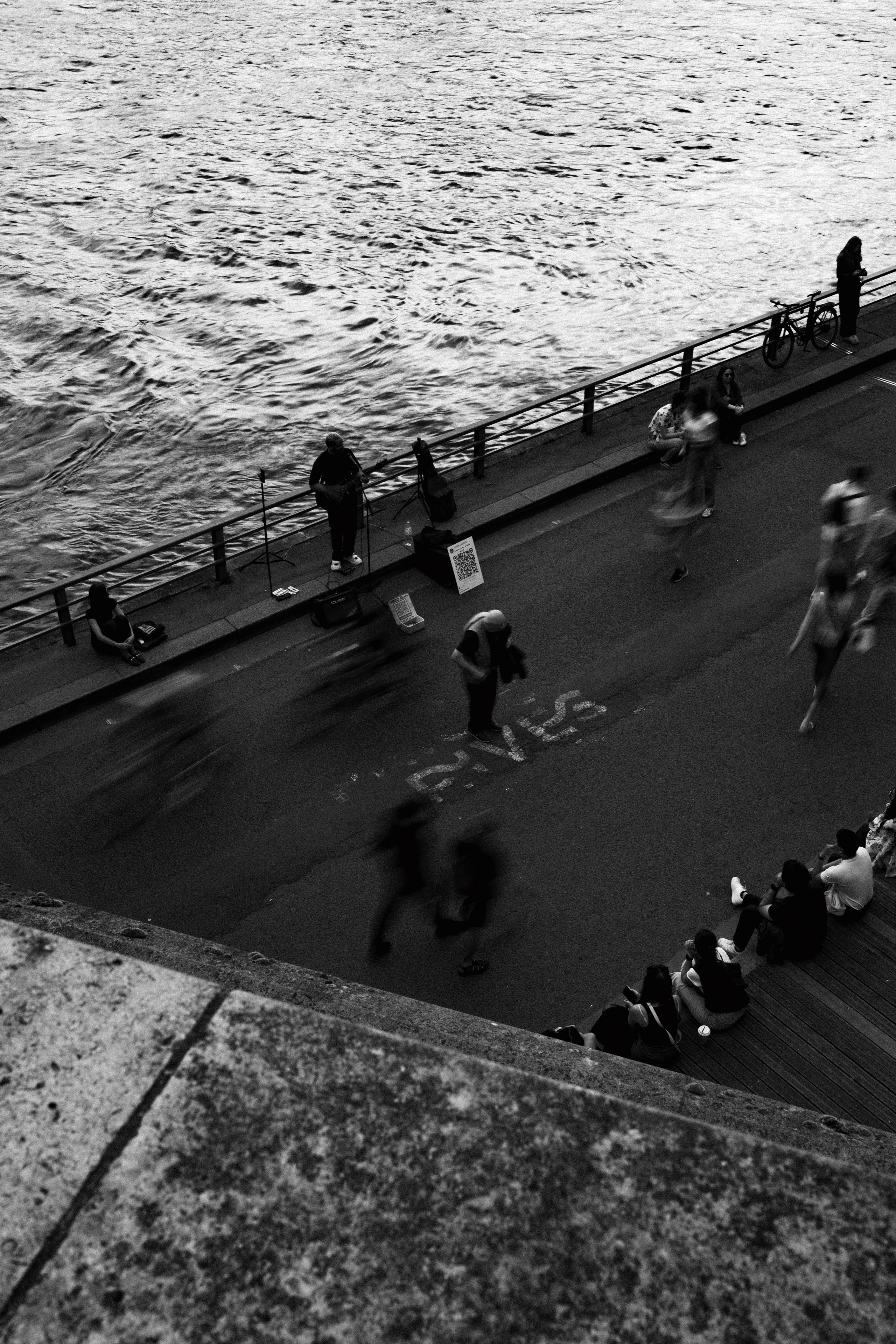 Black and white overhead photo of a riverside promenade in Paris, featuring a street musician playing guitar near the water’s edge, with a QR code sign for tips. Blurred pedestrians and cyclists move swiftly along the designated bike and pedestrian lane marked “CVS.” Onlookers sit on wooden steps near the bottom right, watching or relaxing. The Seine River’s textured water dominates the upper half of the frame, contrasting the stillness of seated figures with the dynamic blur of movement.