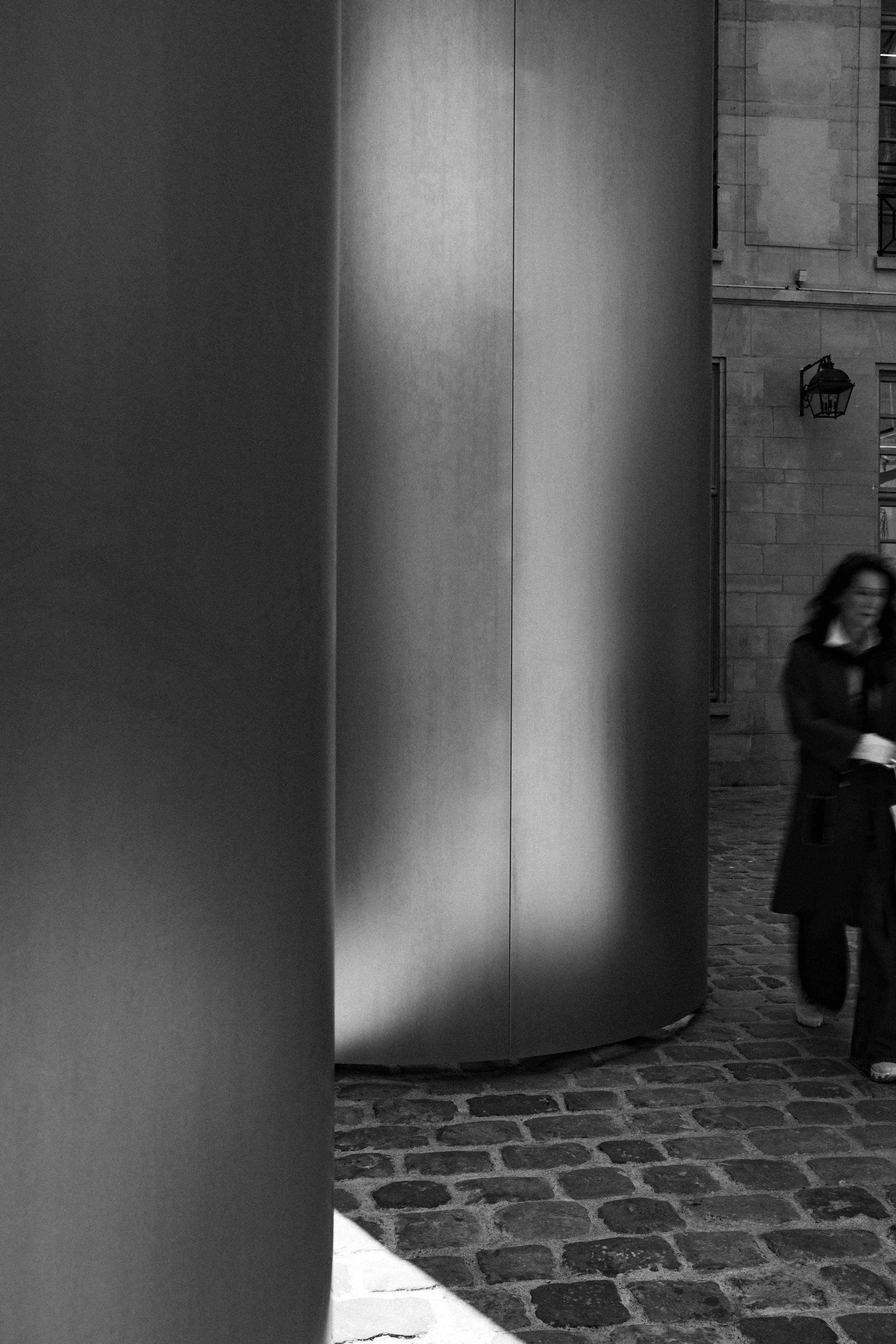 Black and white architectural photograph showcasing a contrast between modern and historic design elements. On the left, large, smooth cylindrical metal columns dominate the composition, reflecting light in soft gradients. To the right, a blurred figure of a woman walks on cobblestone pavement beside a traditional stone building with classic Parisian architectural details, including lantern-style lighting.