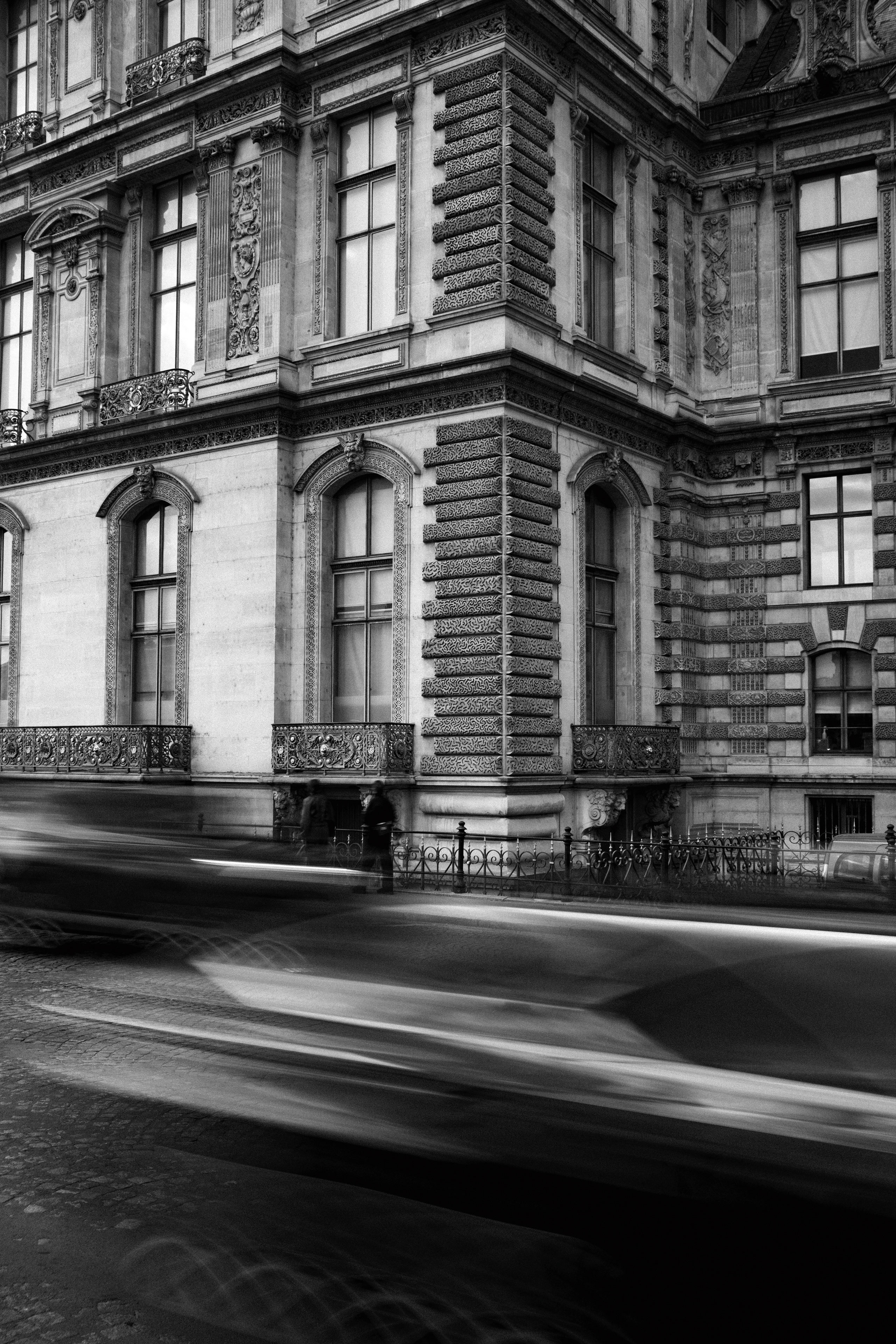 Black and white street photograph capturing motion blur of cars speeding past an ornate, historic Parisian building with elaborate stone carvings, arched windows, and wrought iron balconies. The image combines classical European architecture with dynamic urban energy, emphasized by the streaks of light and shadow from the moving vehicles.