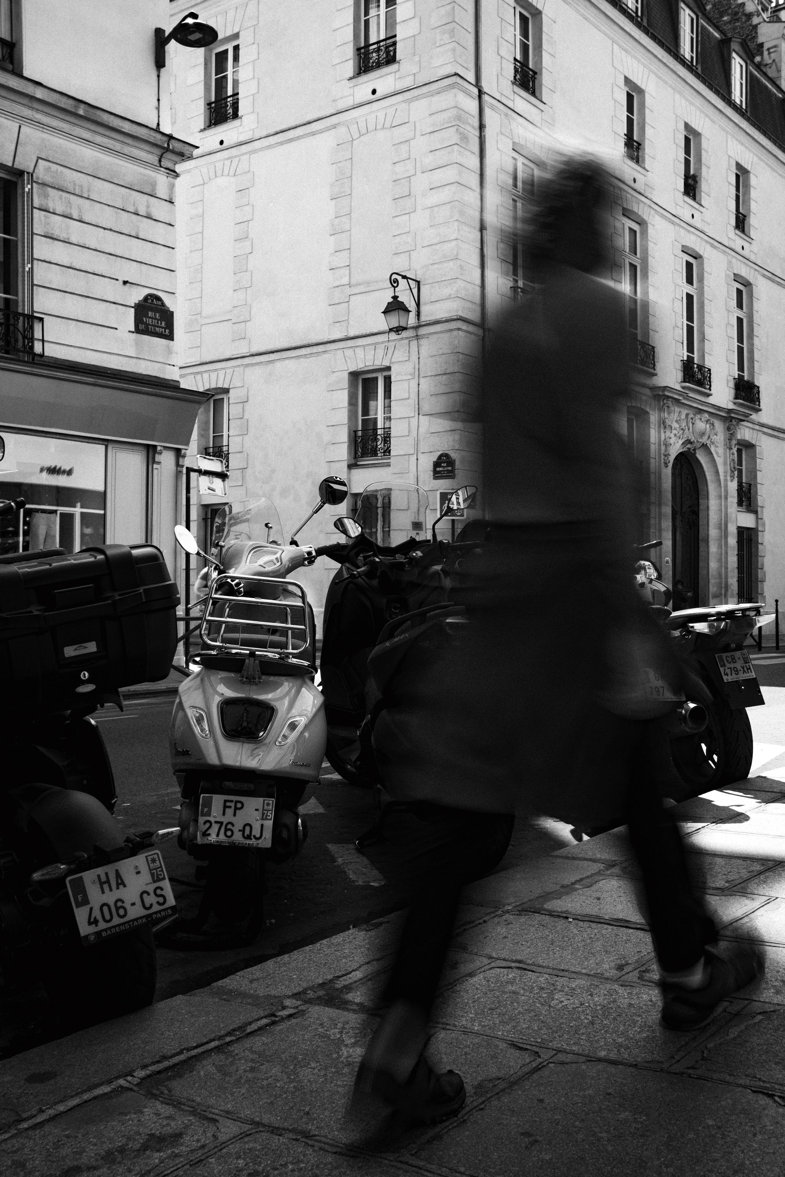 Black and white street photograph capturing a blurred pedestrian walking past a row of parked scooters in front of a classic Parisian corner building. The image is set at the intersection of Rue Vieille du Temple, with visible street signs and traditional French architecture featuring stone facades, wrought iron balconies, and a carved arched doorway. The motion blur of the figure adds dynamic contrast to the stillness of the urban backdrop.