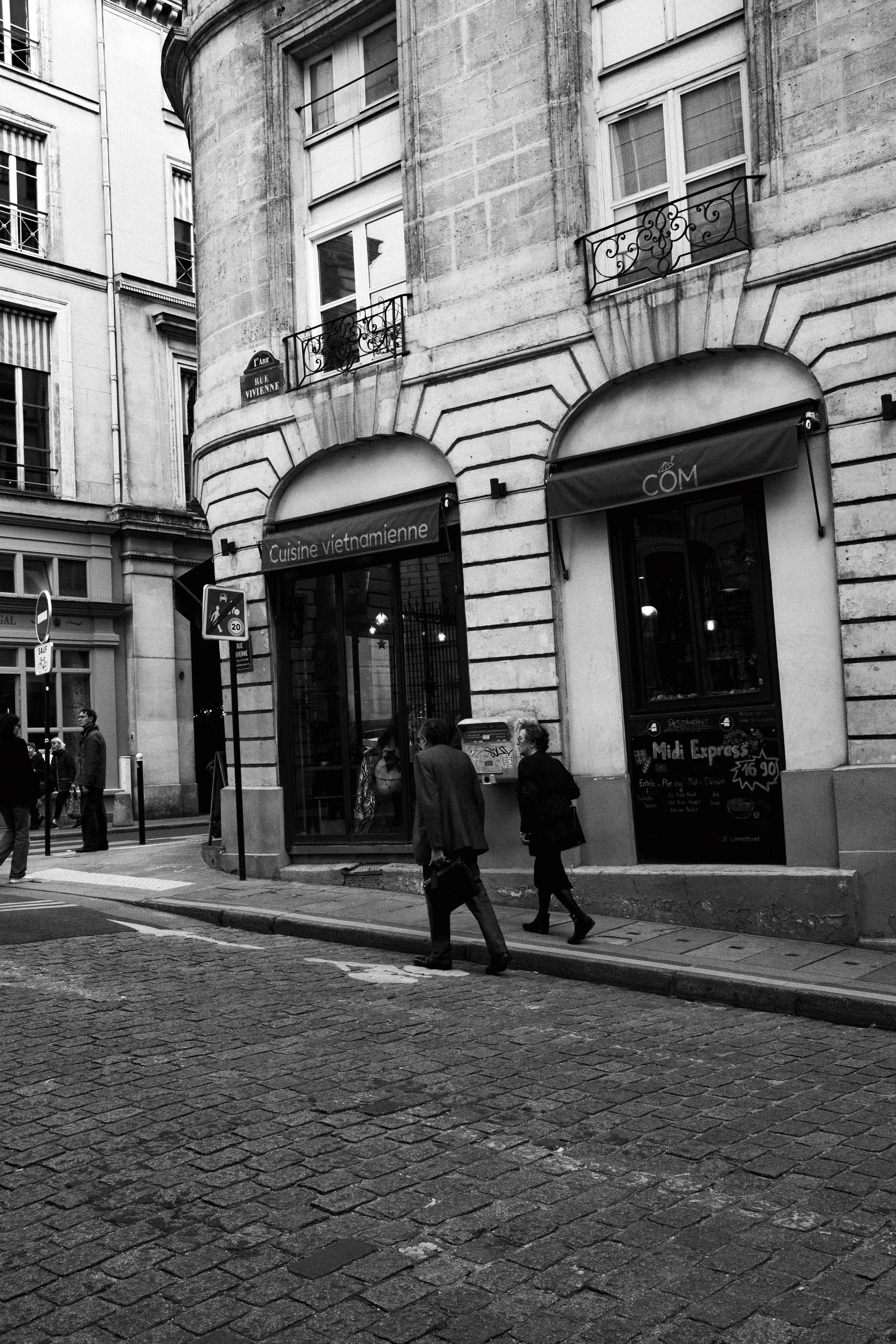 Black and white street photograph capturing a corner view of a traditional European building with arched windows and wrought iron balconies. The ground floor houses a Vietnamese restaurant named “CÔM,” with signage reading “Cuisine vietnamienne” and a chalkboard menu advertising a lunch special for €16.90. Pedestrians walk along the cobblestone street, contributing to the everyday urban atmosphere. The street signs and architecture suggest a French city setting.