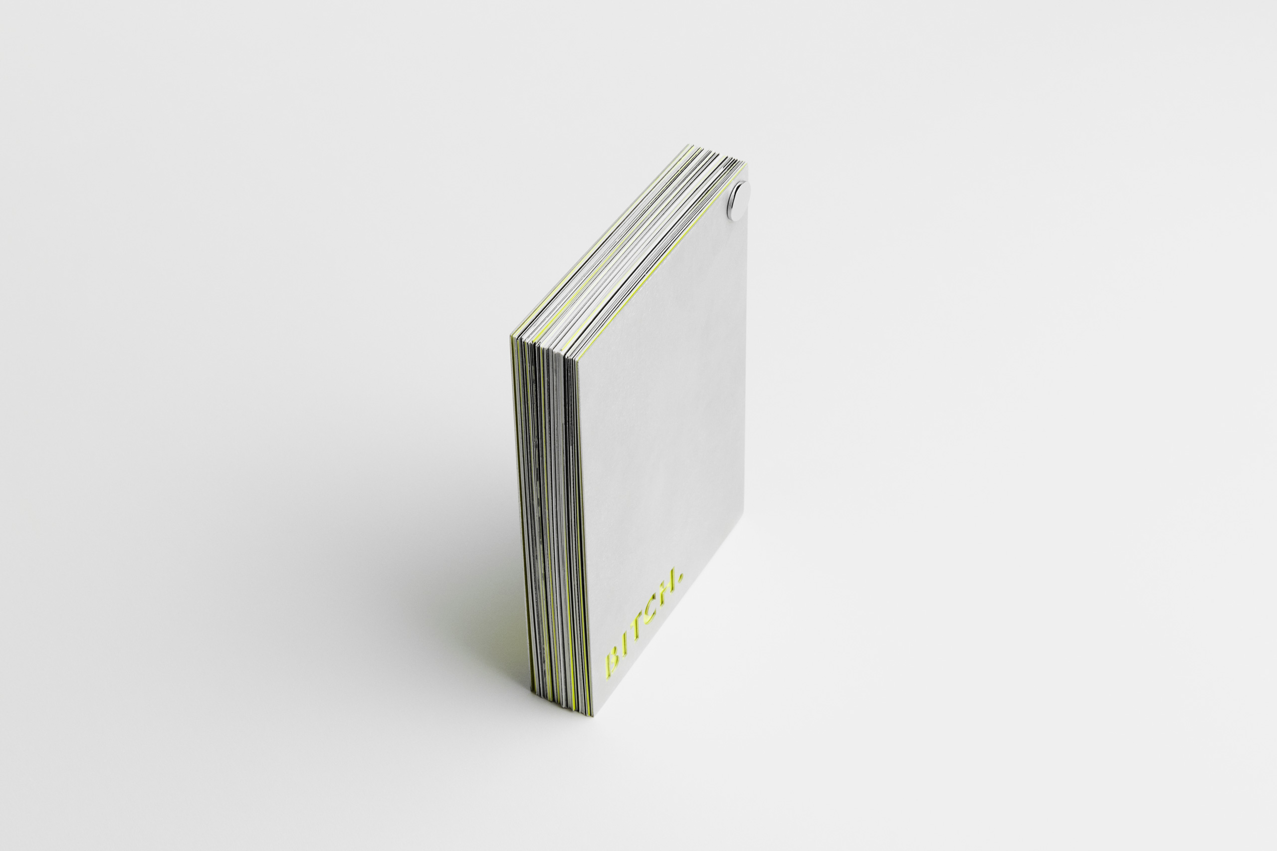 Vertical angled view of a thick, modern flipbook with a matte gray cover, secured by a metal screw post in the top-right corner. The front cover features bold neon yellow text “BITCH.” near the bottom left edge. The edges of the inner pages reveal a layered color scheme of white, gray, black, and neon yellow, emphasizing a high-contrast, edgy design.