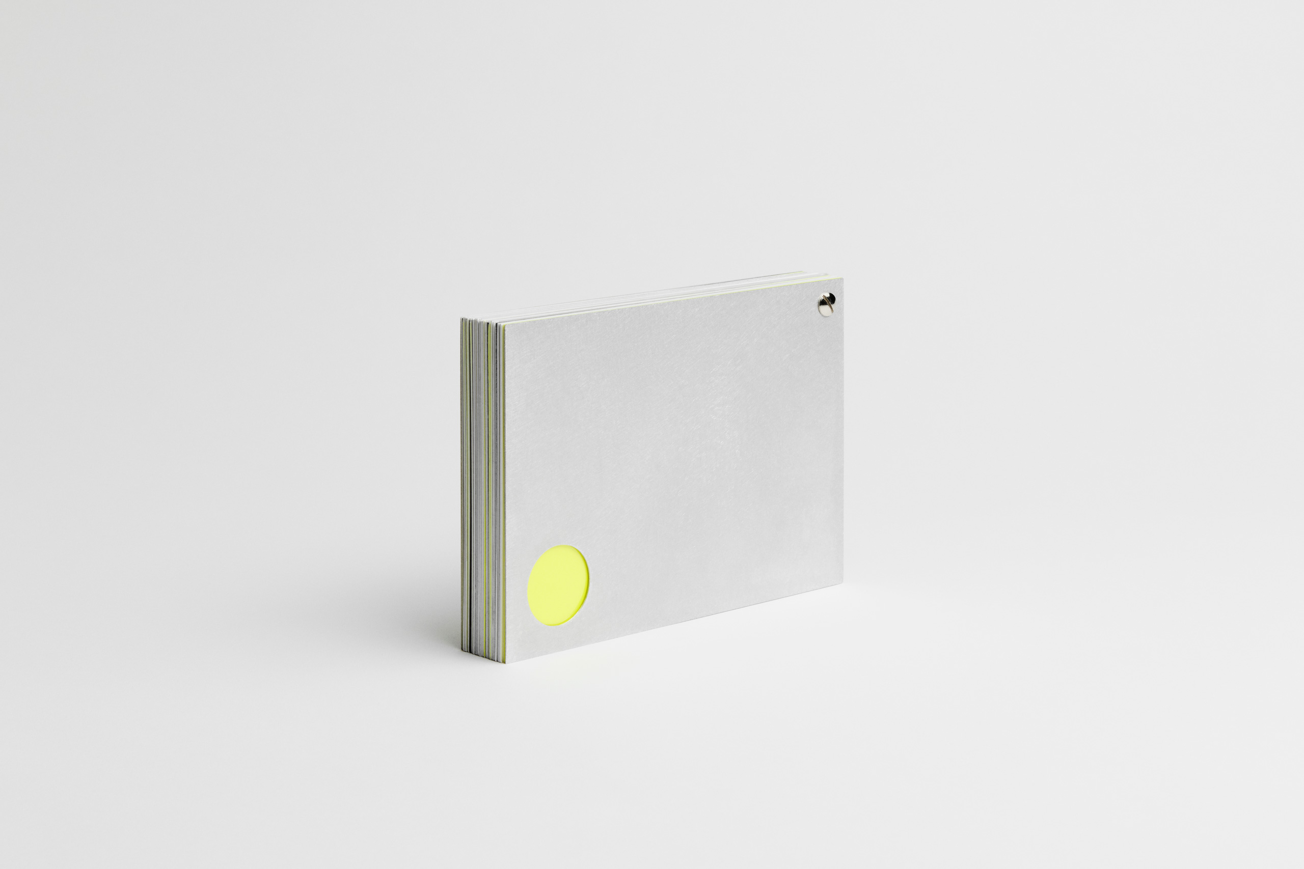 Angled side view of a thick, horizontally oriented gray booklet with a metallic screw post binding at the top-right and a circular cut-out near the bottom-left revealing a neon yellow page beneath. The layered pages show subtle neon yellow and grayscale edge details, emphasizing a bold yet minimalist design.