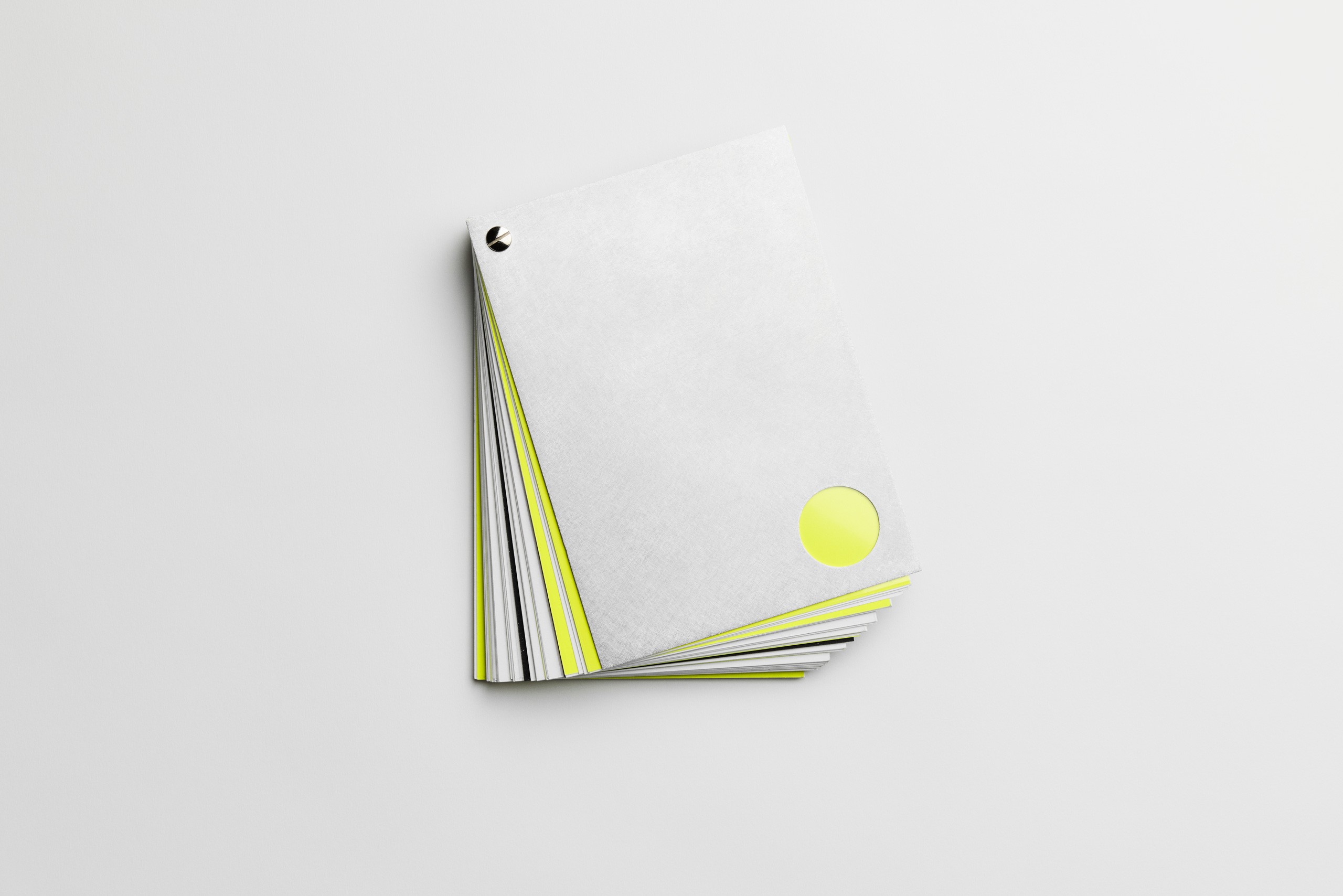 Angled view of a partially fanned-out minimalist booklet with a matte gray cover, featuring a metallic screw post binding in the top-left corner and a circular cut-out at the bottom-right that reveals a neon yellow inner page. The visible interior pages show alternating gray, white, and neon yellow edges, enhancing the visual contrast.