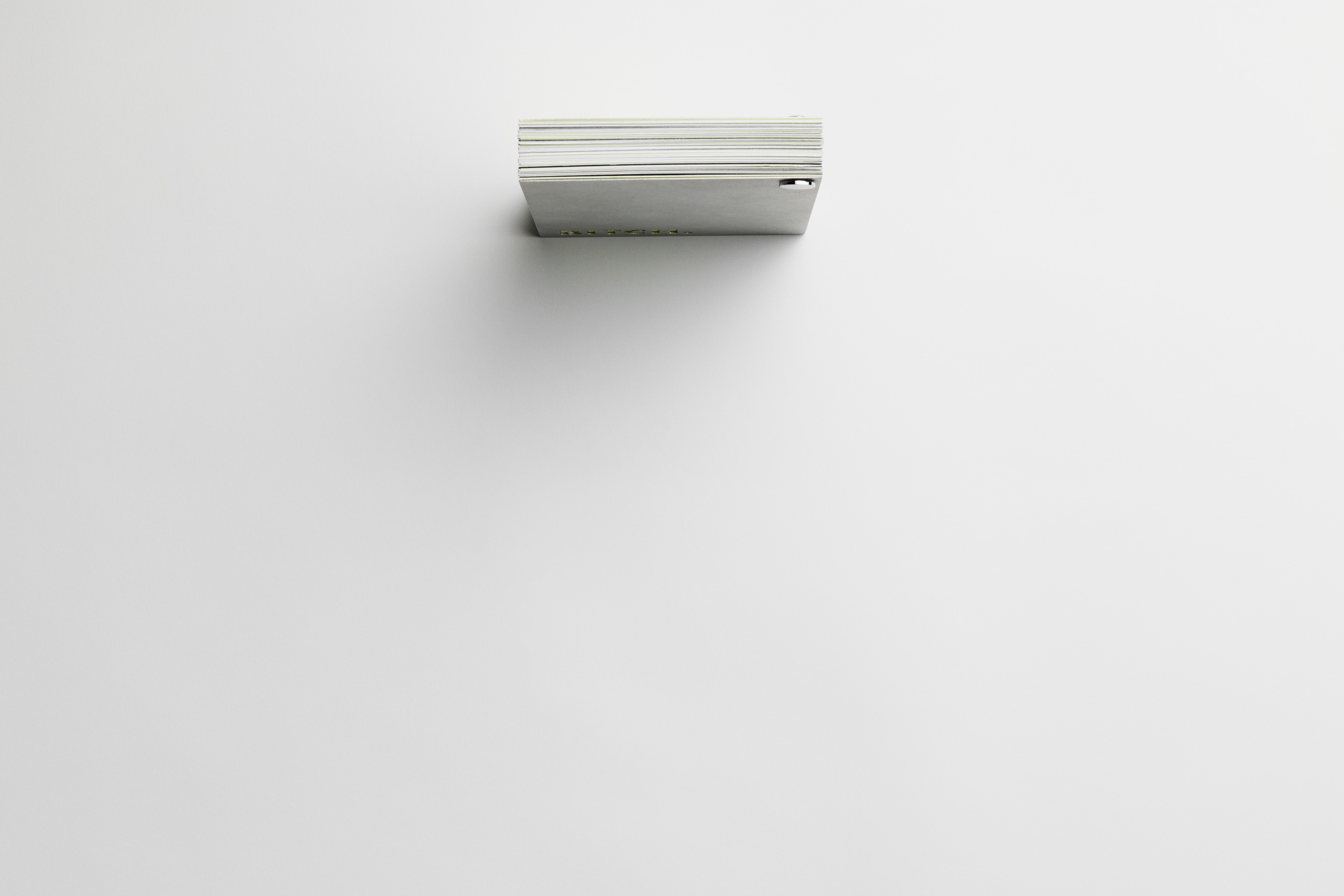Top-down view of a closed, horizontally positioned booklet with a thick stack of pages, bound by a metallic fastener. The visible spine reveals layered pages in neutral tones with subtle hints of neon yellow. The front cover, faintly displaying the word “BITCH.” in neon yellow, contributes to the bold, minimalist aesthetic.