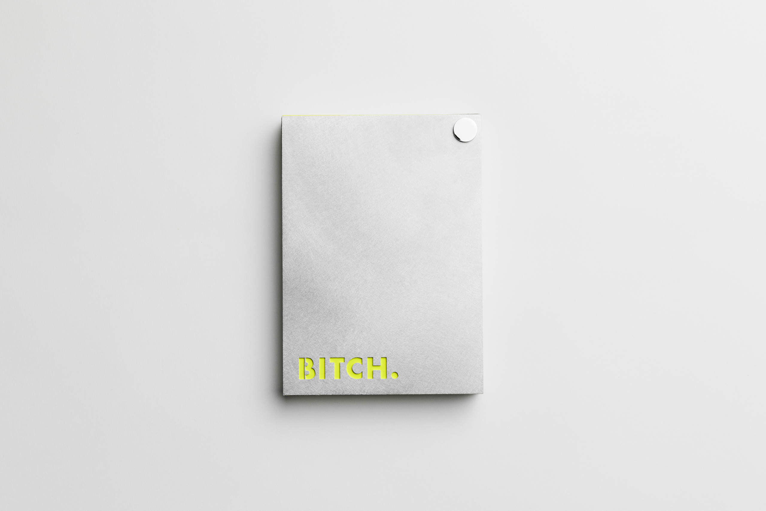 Minimalist gray notebook on a white background, featuring a bold neon yellow word “BITCH.” in all caps at the bottom left corner; a small round white sticker or magnet is affixed to the top right corner. This image is styled with clean, modern aesthetics and uses provocative typography for edgy branding or artistic statement.