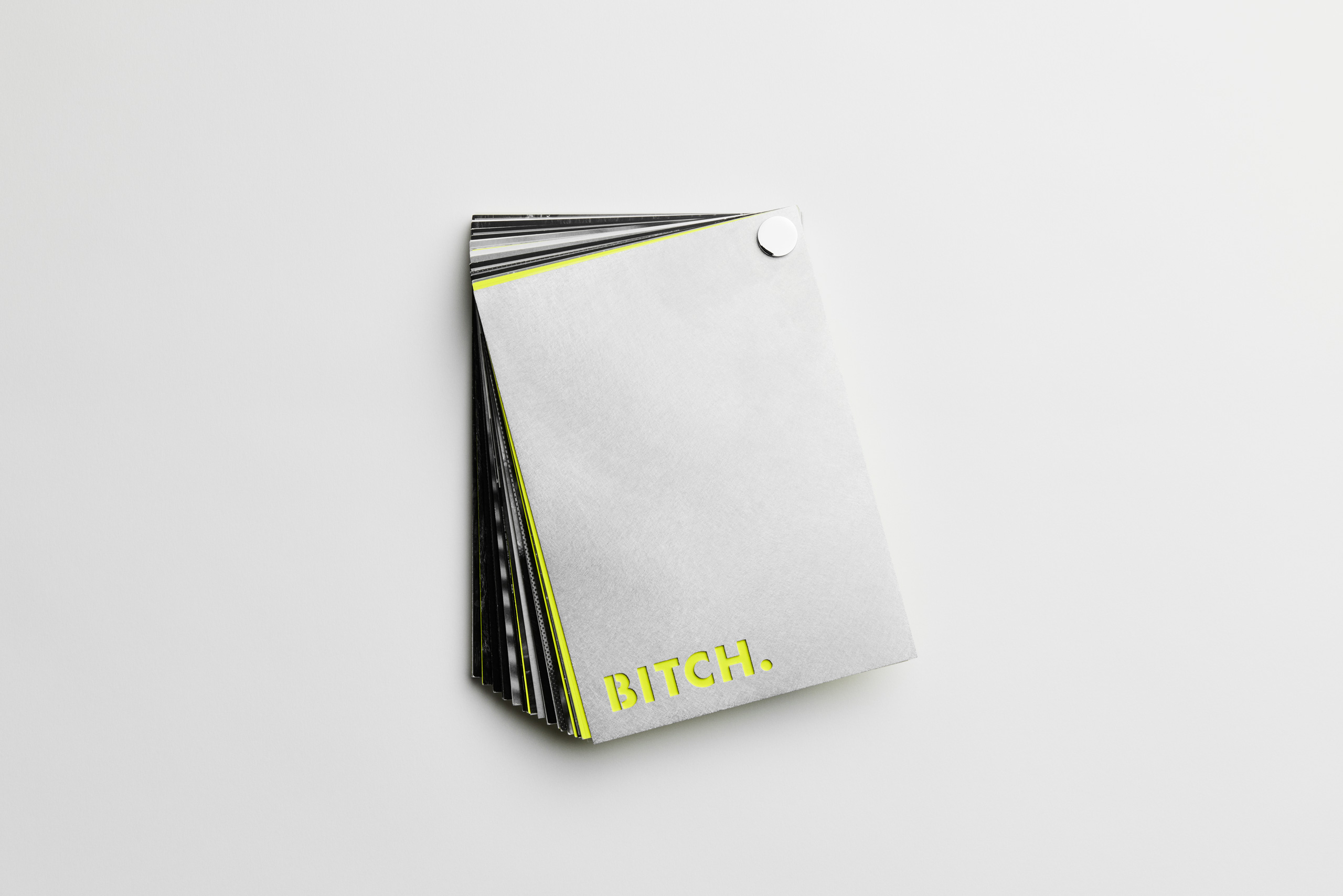 Angled view of a modern gray flipbook with slightly fanned-out pages, secured by a white metal fastener in the top-right corner; the cover features bold neon yellow text “BITCH.” in all caps at the bottom left. Inner pages have visible black and neon accents, suggesting a high-contrast, graphic design theme.