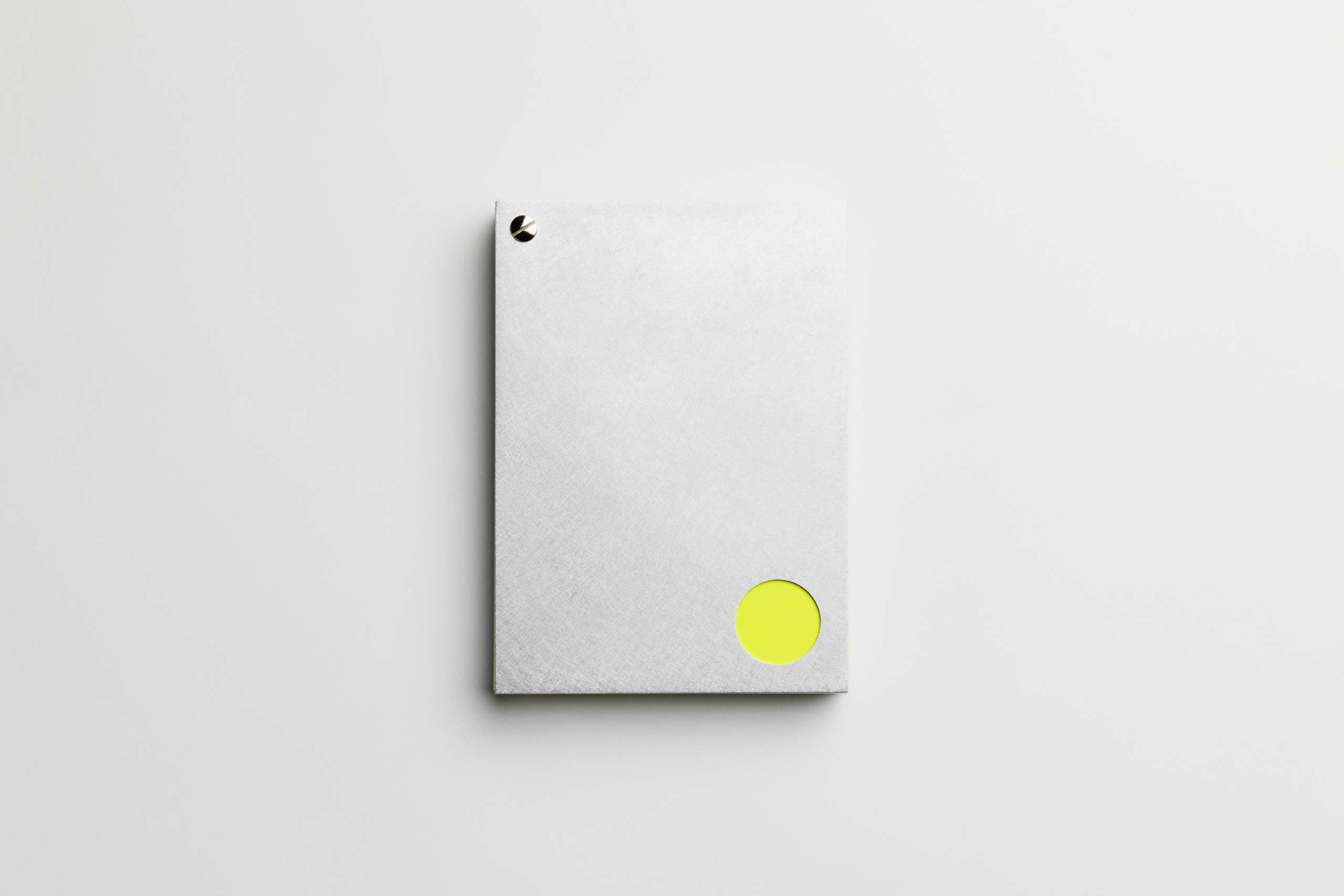Minimalist gray notebook or booklet with a silver binding rivet in the top-left corner and a circular cut-out at the bottom-right corner revealing a neon yellow circle beneath. The design is clean and modern, emphasizing contrast between neutral tones and vibrant color.