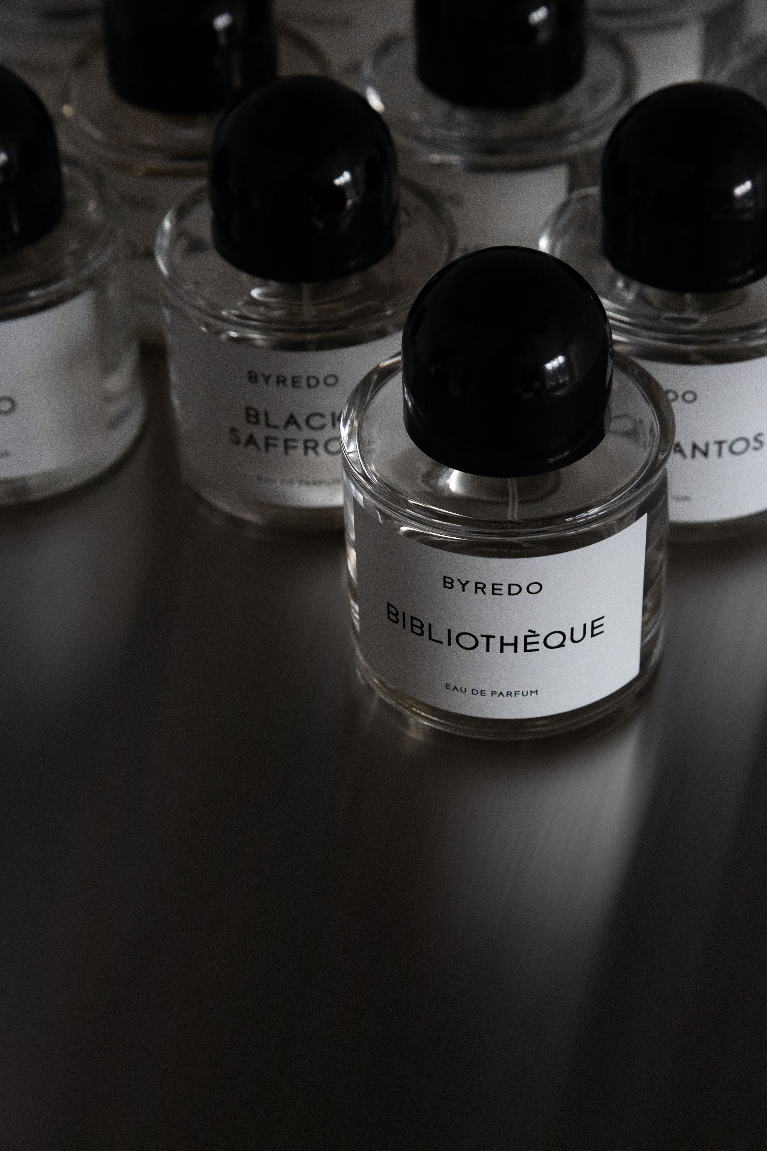 Close-up photo of multiple Byredo perfume bottles, with the central focus on a clear glass bottle labeled “BYREDO BIBLIOTHÈQUE Eau de Parfum” featuring a minimalist white label and black cap. Surrounding bottles include other fragrances like “Black Saffron” and “Santos,” arranged on a sleek, reflective dark surface. Luxury fragrance collection, niche perfume branding, high-end scent packaging, and designer eau de parfum photography.