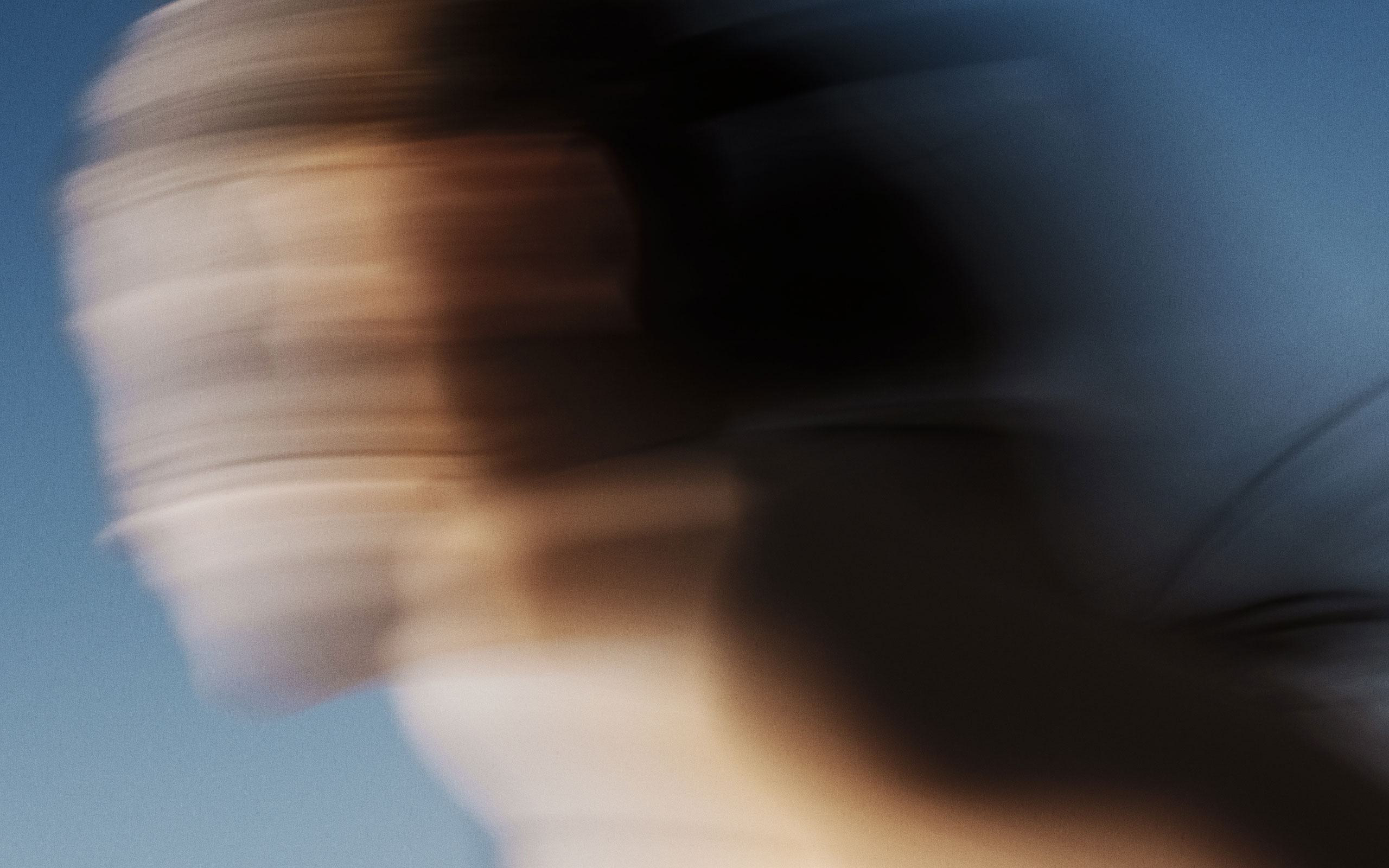 Abstract motion-blur photograph of a person running, with warm skin tones and dark shadows blending against a clear blue sky background. The image conveys dynamic movement, speed, and energy, creating a visually artistic representation of athleticism and outdoor activity.