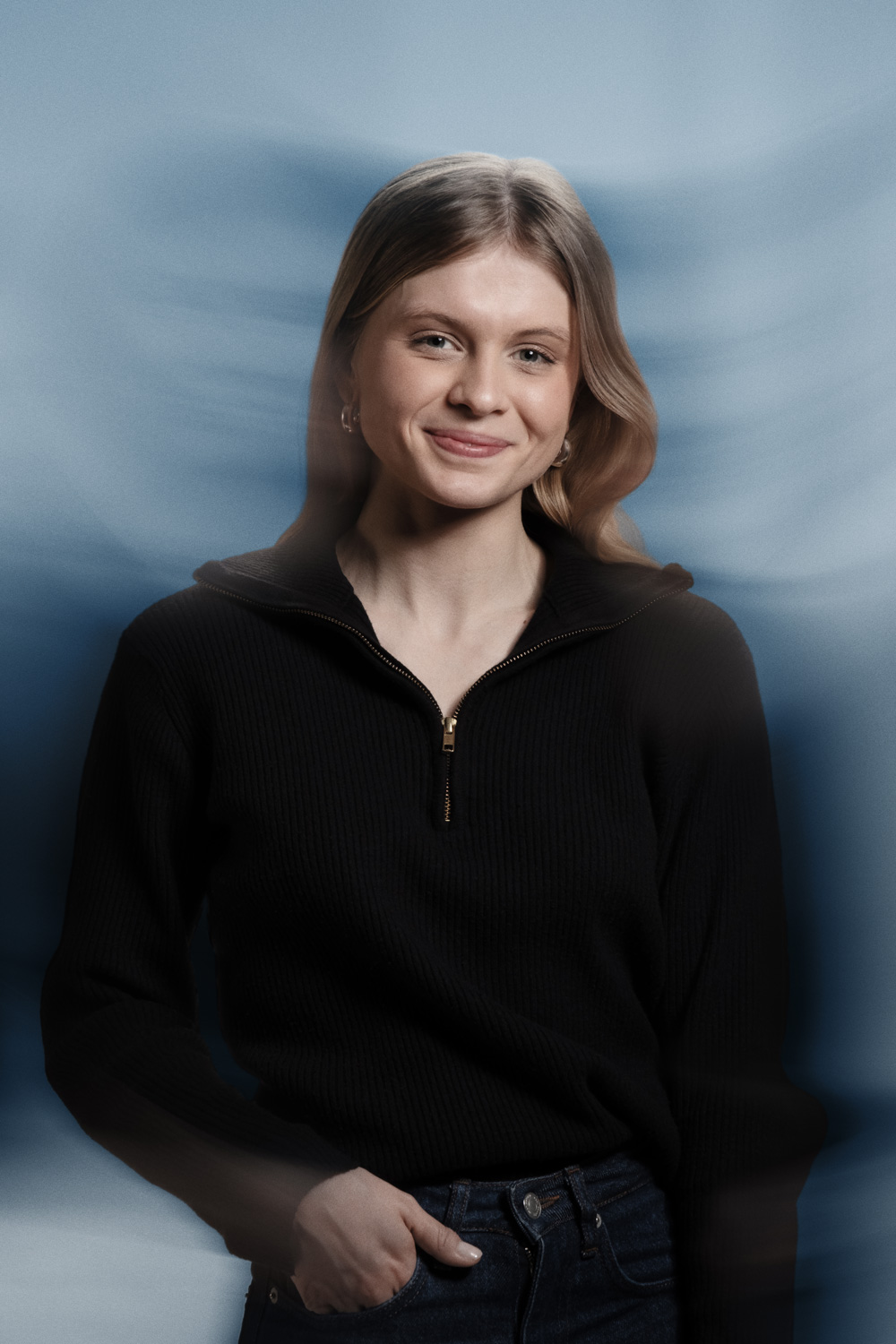 Portrait of a young woman with long blonde hair smiling softly against a soft blue background with subtle motion blur effects. She is wearing a black ribbed zip-collar sweater and dark jeans, with one hand in her pocket, exuding a calm and confident demeanor. The image features a modern, stylized aesthetic ideal for professional profile pictures, branding photography, or creative headshots.