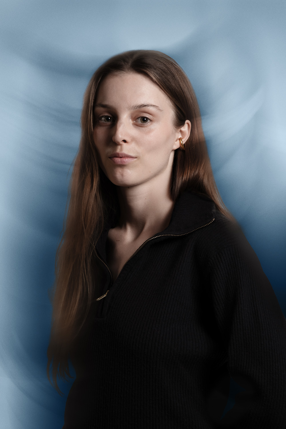 Portrait of a young woman with long straight brown hair and a neutral expression, posed against a light blue background with soft motion blur effects. She is wearing a black ribbed zip-up sweater with a gold zipper and gold hoop earrings, presenting a clean, modern, and composed aesthetic. Ideal for editorial portraits, creative headshots, or branding imagery.