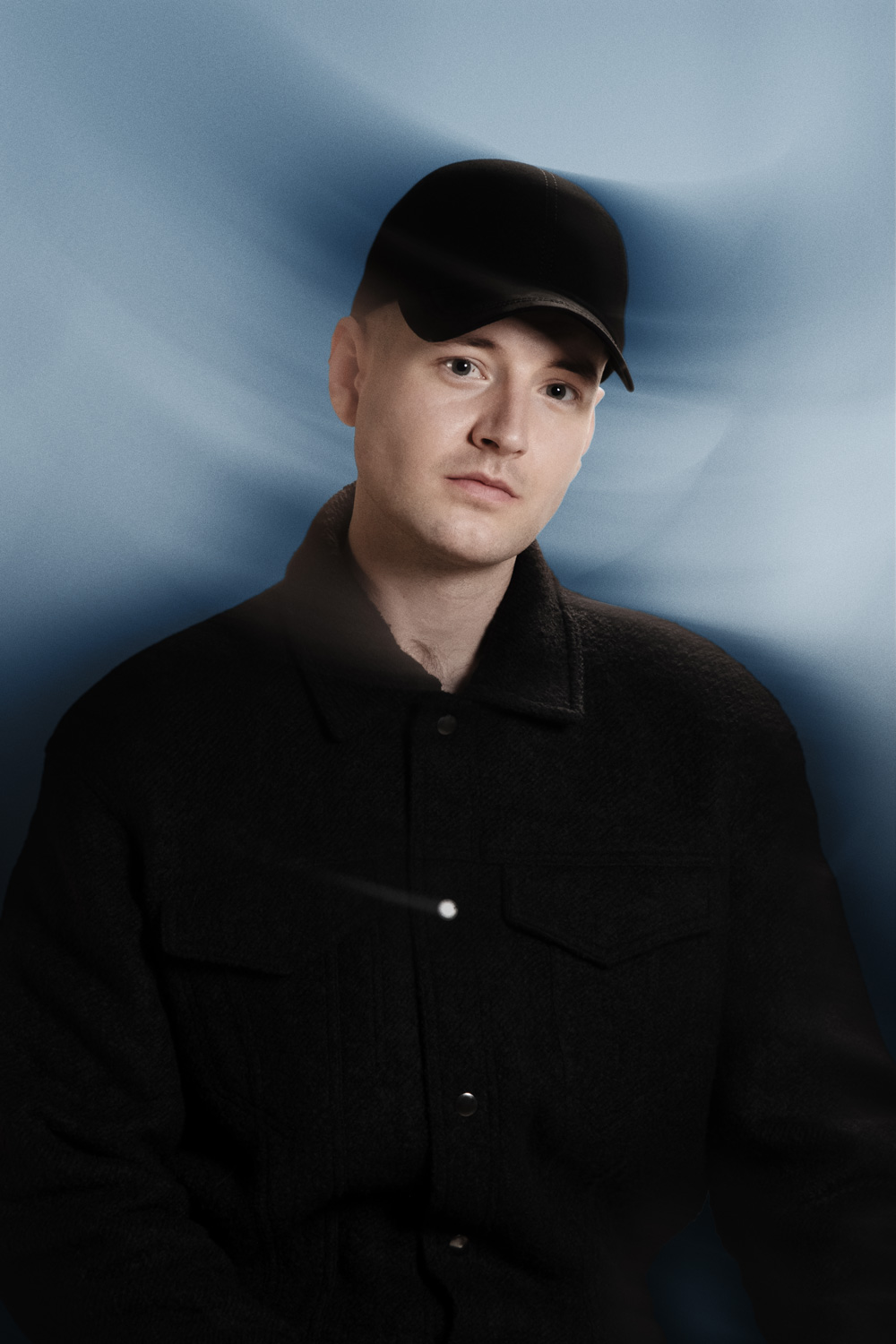 Portrait of a young man wearing a black baseball cap and a black wool jacket, posed against a light blue background with soft motion blur accents. He has a calm, serious expression, and the photo features a minimalist, modern aesthetic with moody lighting and a stylized atmosphere.