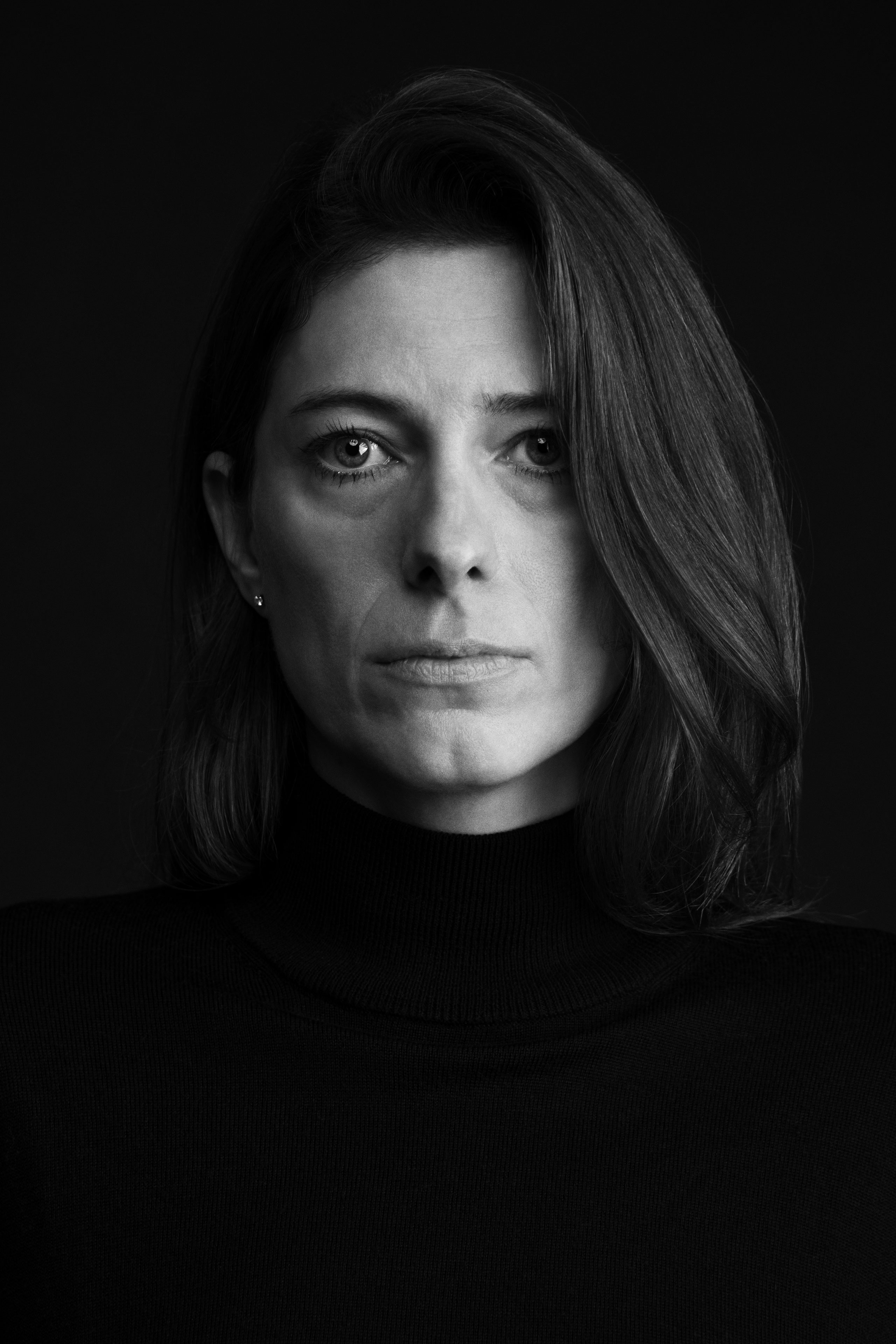 Black and white portrait of a woman with shoulder-length hair parted to the side, wearing a black turtleneck sweater, looking directly at the camera with a calm yet serious expression against a solid dark background.