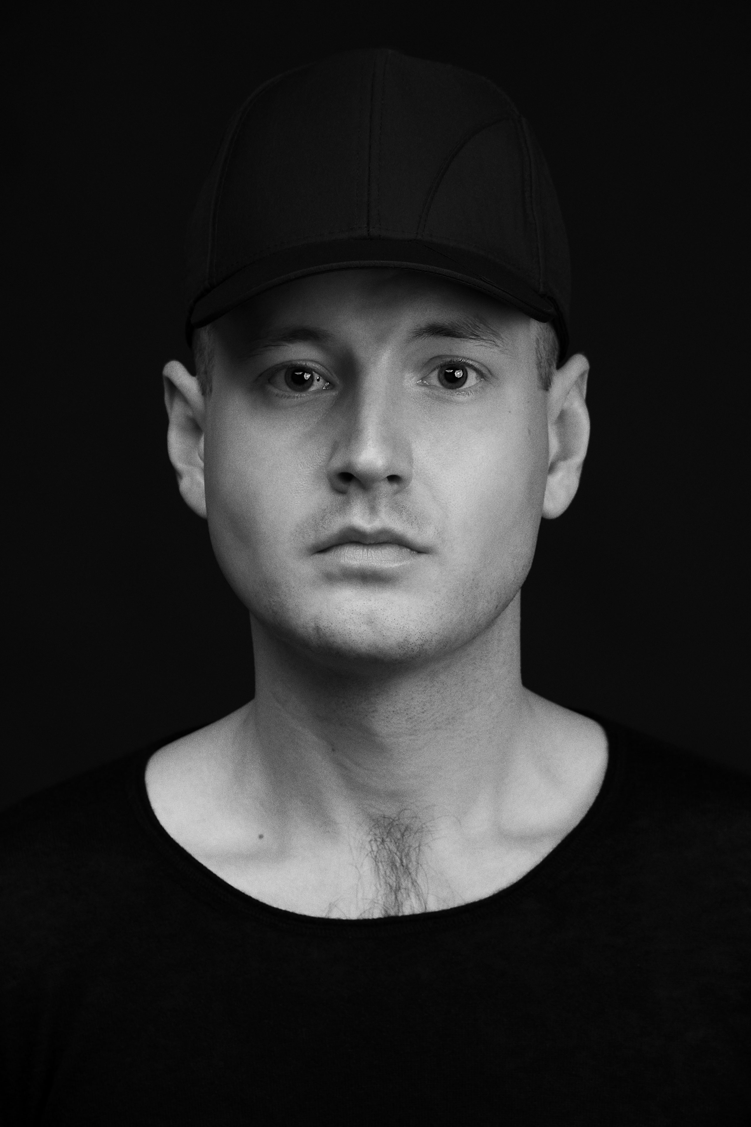 Black and white portrait of a man wearing a plain black baseball cap and a wide neckline black shirt, looking directly at the camera with a neutral, composed expression against a solid dark background.