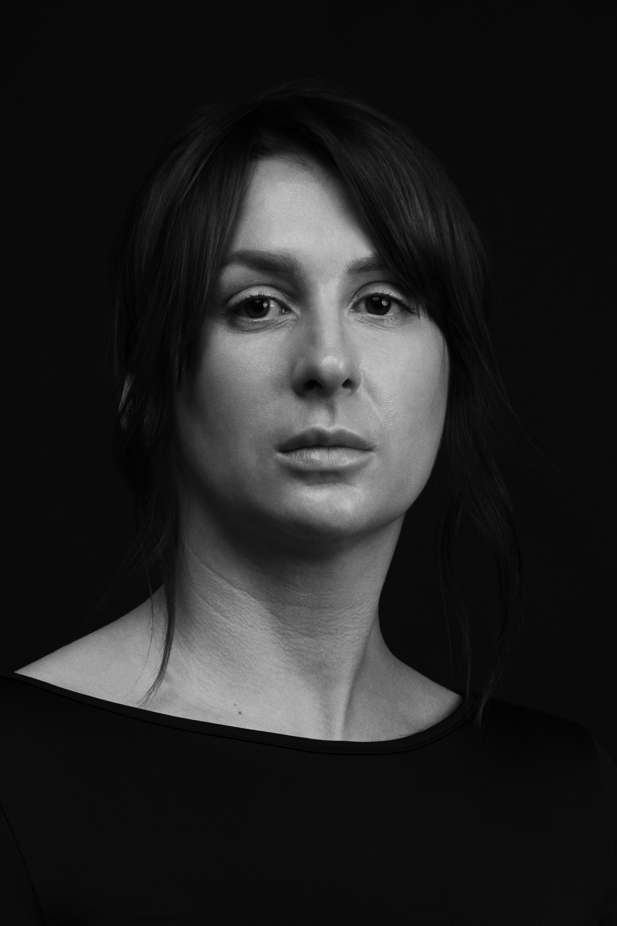 Black and white portrait of a woman with dark hair styled in a loose, low updo with soft strands framing her face, wearing a wide neckline black top, gazing directly at the camera with a composed, neutral expression against a solid dark background.