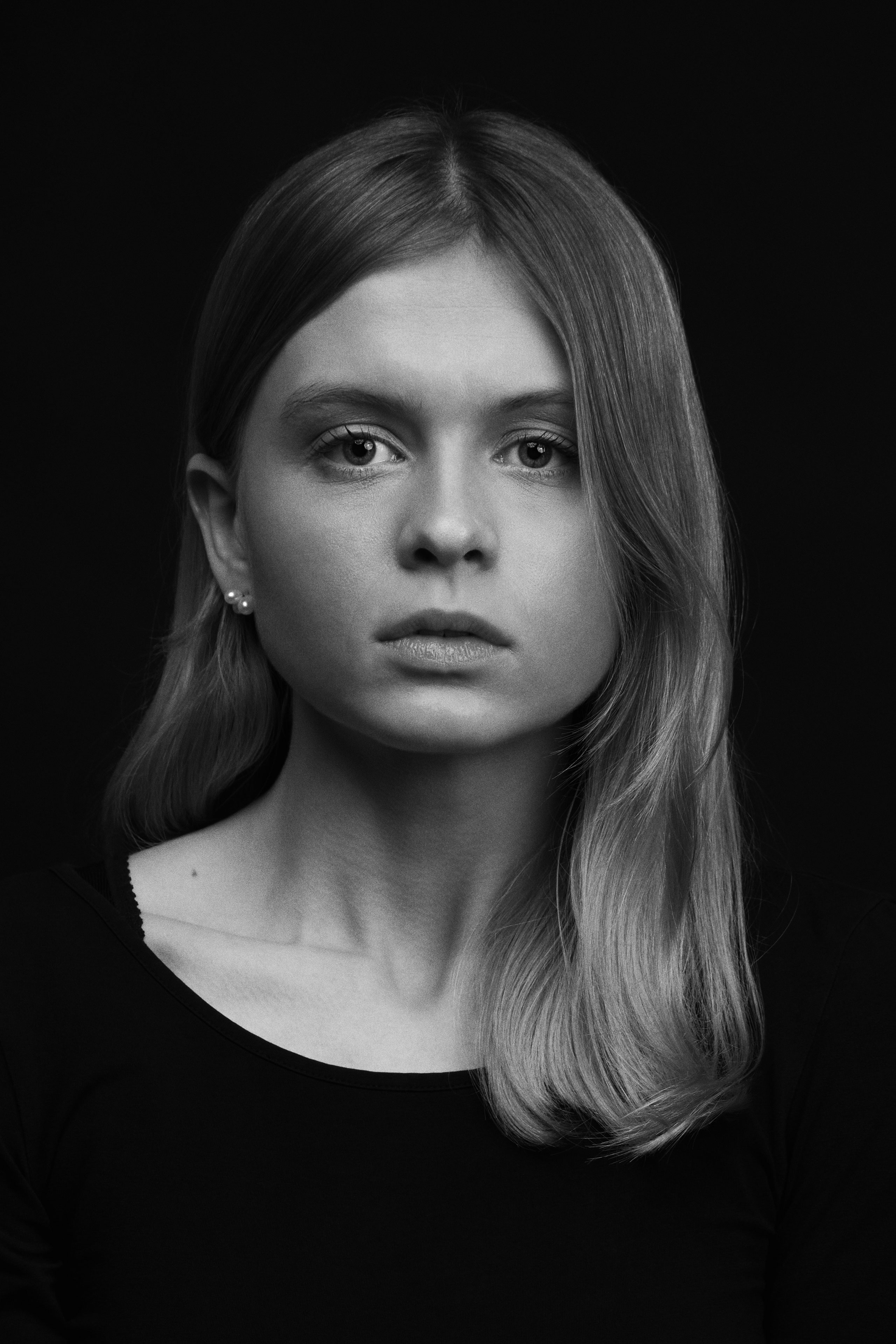 Black and white portrait of a young woman with straight, shoulder-length hair parted slightly off-center, wearing a plain black top and small stud earrings, gazing directly at the camera with a serious, neutral expression against a solid dark background.