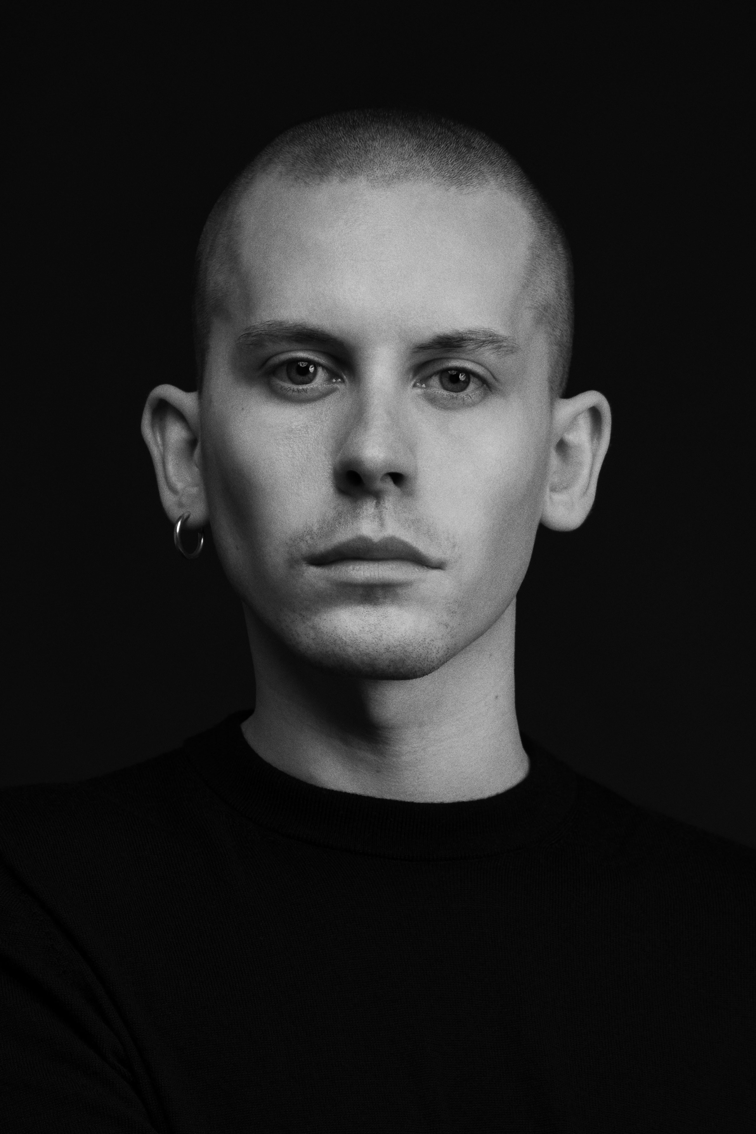 Black and white portrait of a young person with a shaved head, wearing a plain black crewneck sweater and a small hoop earring in the left ear, looking directly at the camera with a neutral expression against a solid dark background.